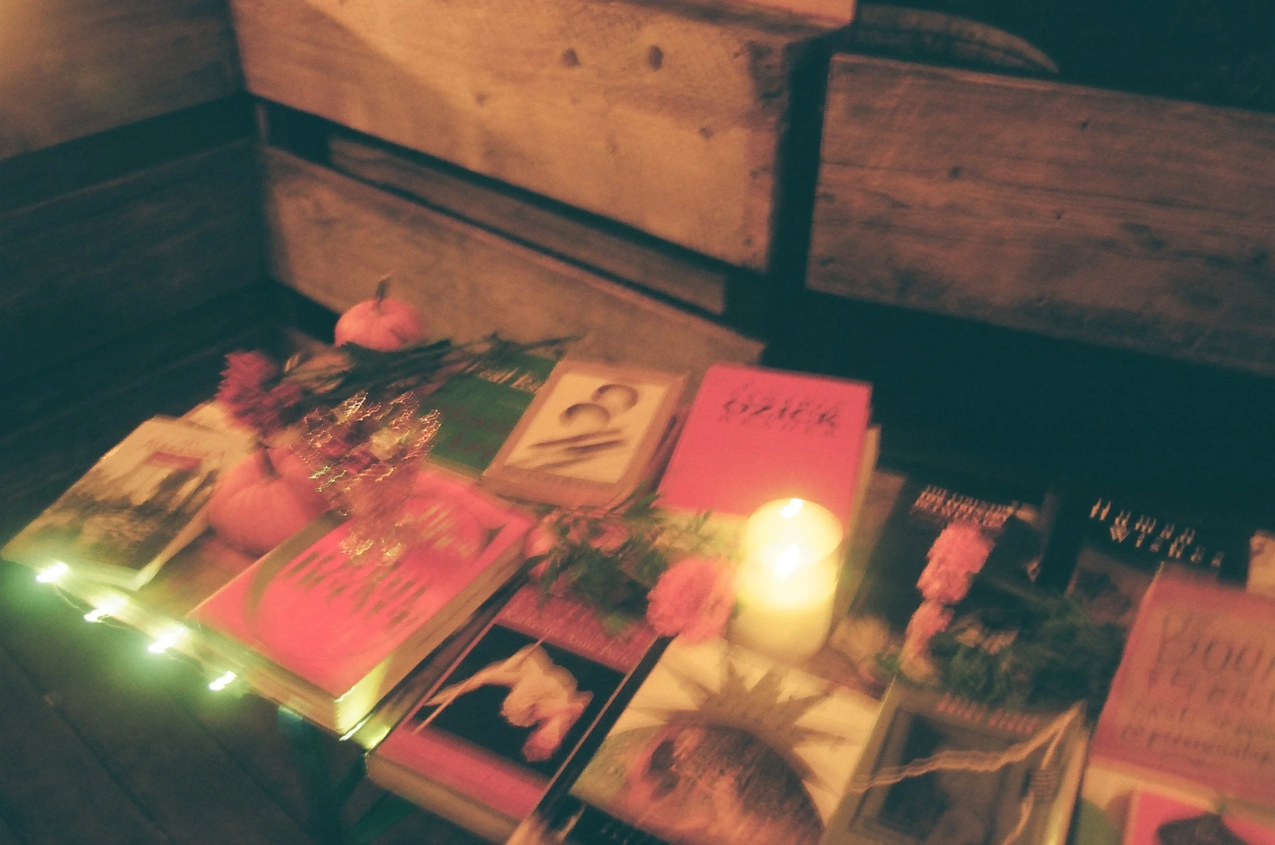 Books and a lit candle on a table, with pink flowers and a small pumpkin in the background.