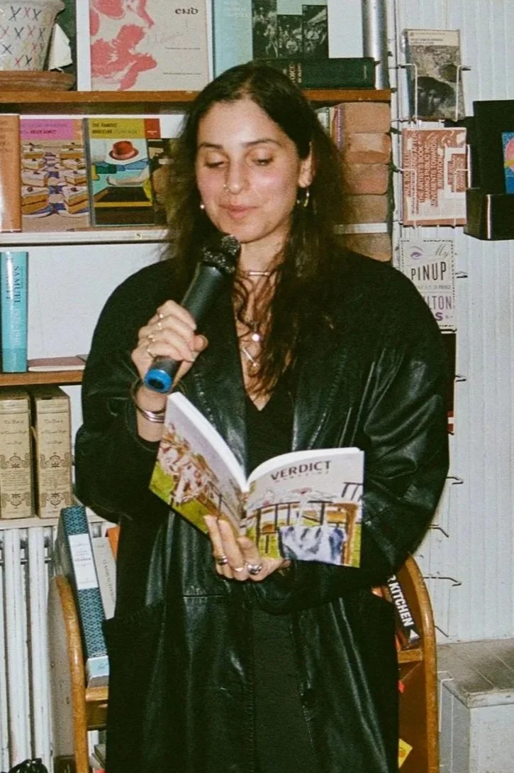 A woman with dark hair, wearing a black leather jacket and holding a microphone and a small book titled "VERDICT," appears to be speaking or reading in a bookstore or library with shelves of books behind her.