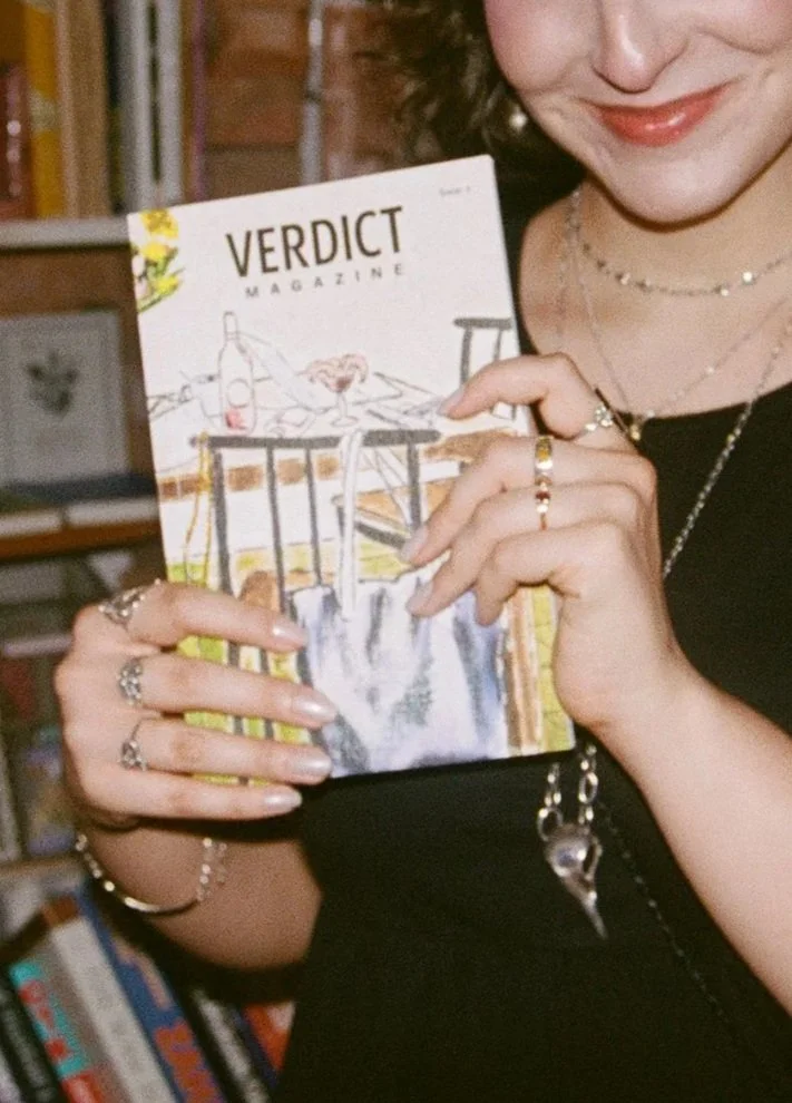 A woman holding a magazine titled 'Verdict Magazine', her face partially visible, wearing jewelry, in a room with bookshelves in the background.