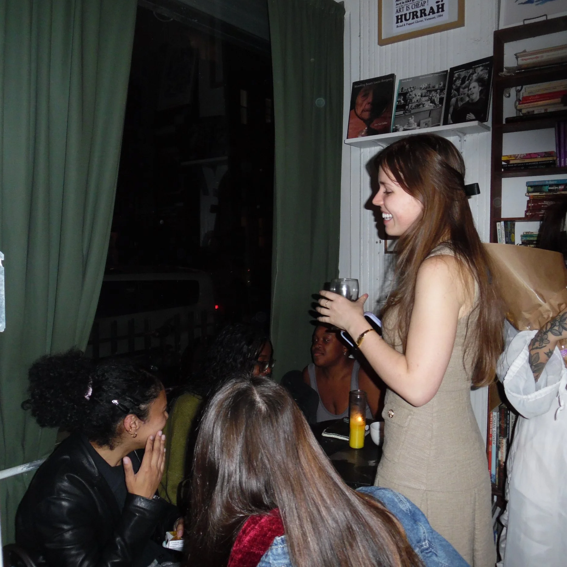 A woman with long brown hair in a beige dress holds a drink and smiles while speaking to a group of women sitting at a table inside a cozy room with bookshelves and framed photos on the wall.