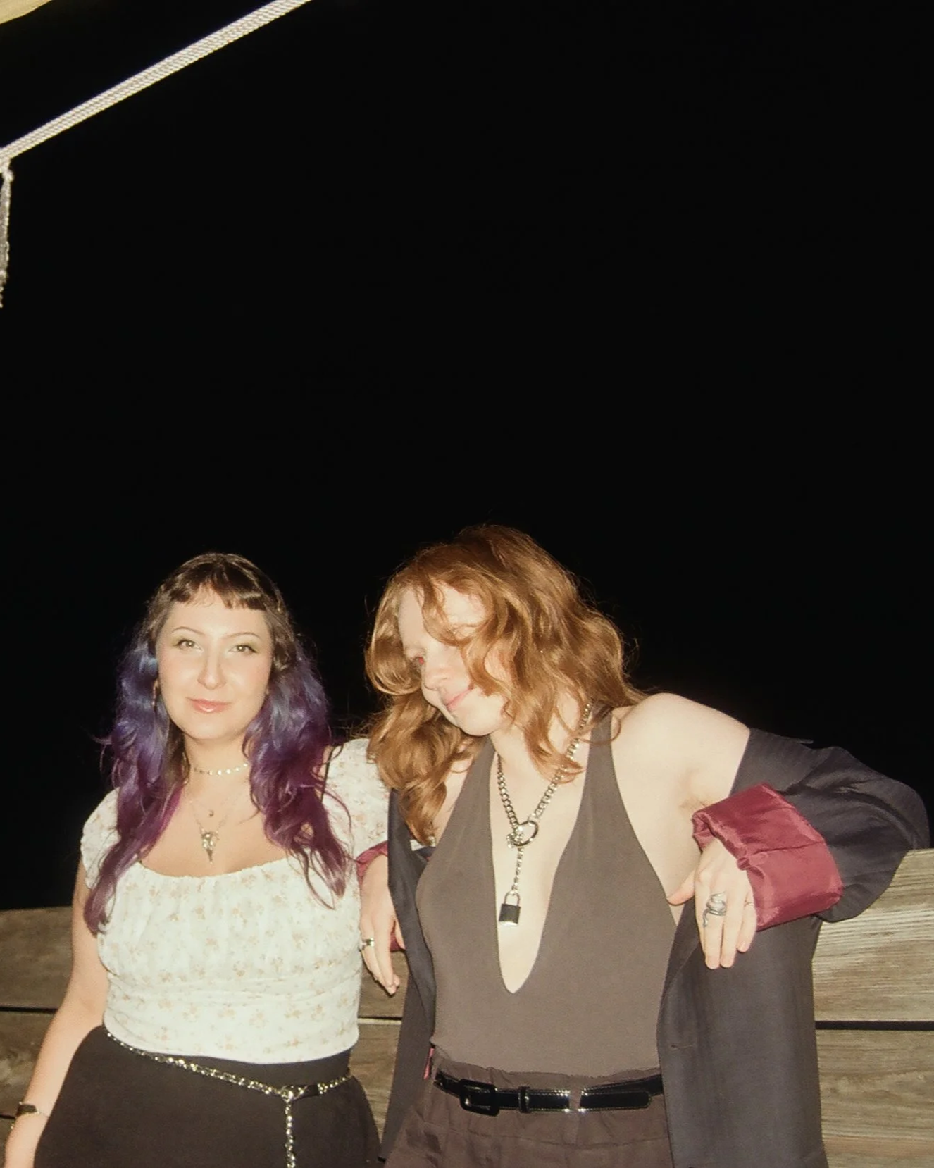 Two women standing outdoors at night, one with purple-tinted hair and the other with red hair, posing casually.
