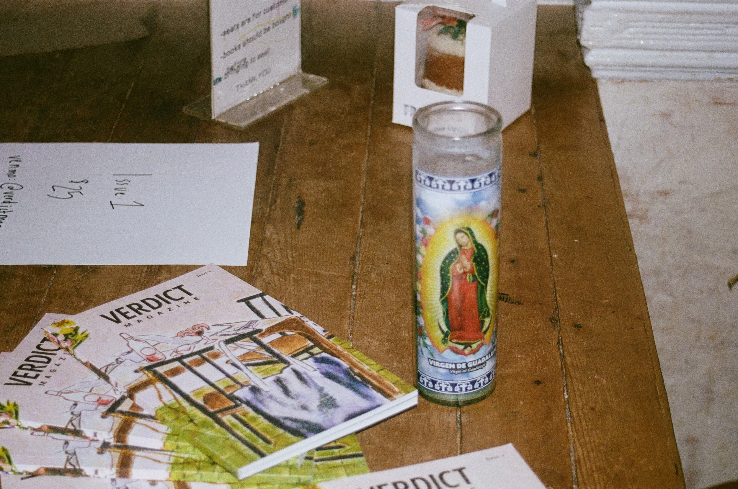 A table with religious items including a candle with an image of the Virgin of Guadalupe, several copies of Veridict magazine, and handwritten notes, with some flyers and boxes nearby.