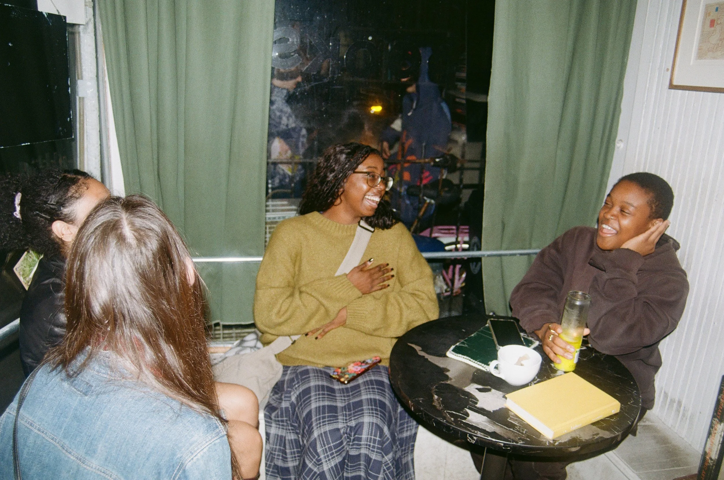 A group of five people sitting around a black marble table, smiling and chatting inside a cozy cafe, with one person holding a drink, and a phone, notebook, and cup on the table.