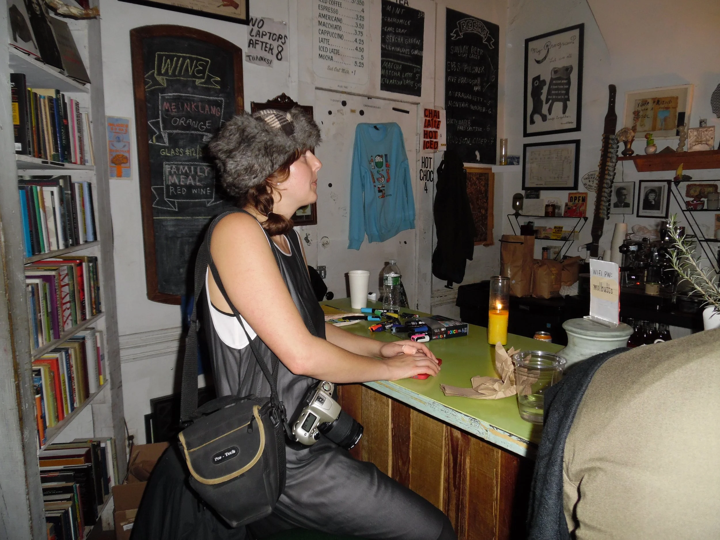 A woman with a camera hanging from her neck and a furry hat, sitting at a green counter in a cozy cafe, with bookshelves on the left and various framed pictures and signs on the wall behind her.