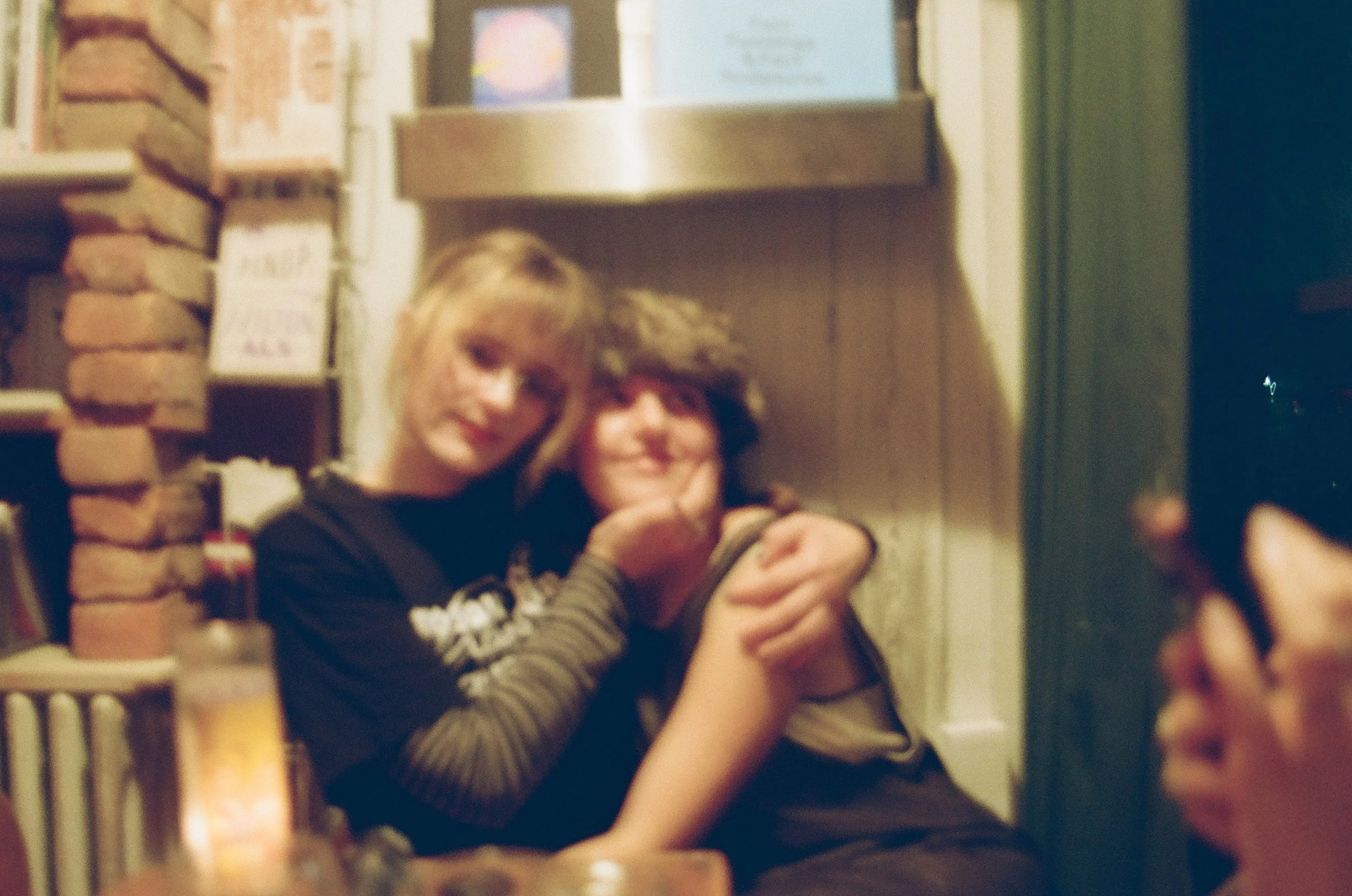 Two children sitting close together, one with blond hair and the other with brown hair, in a cozy room with brick and wood walls, smiling and in close embrace.