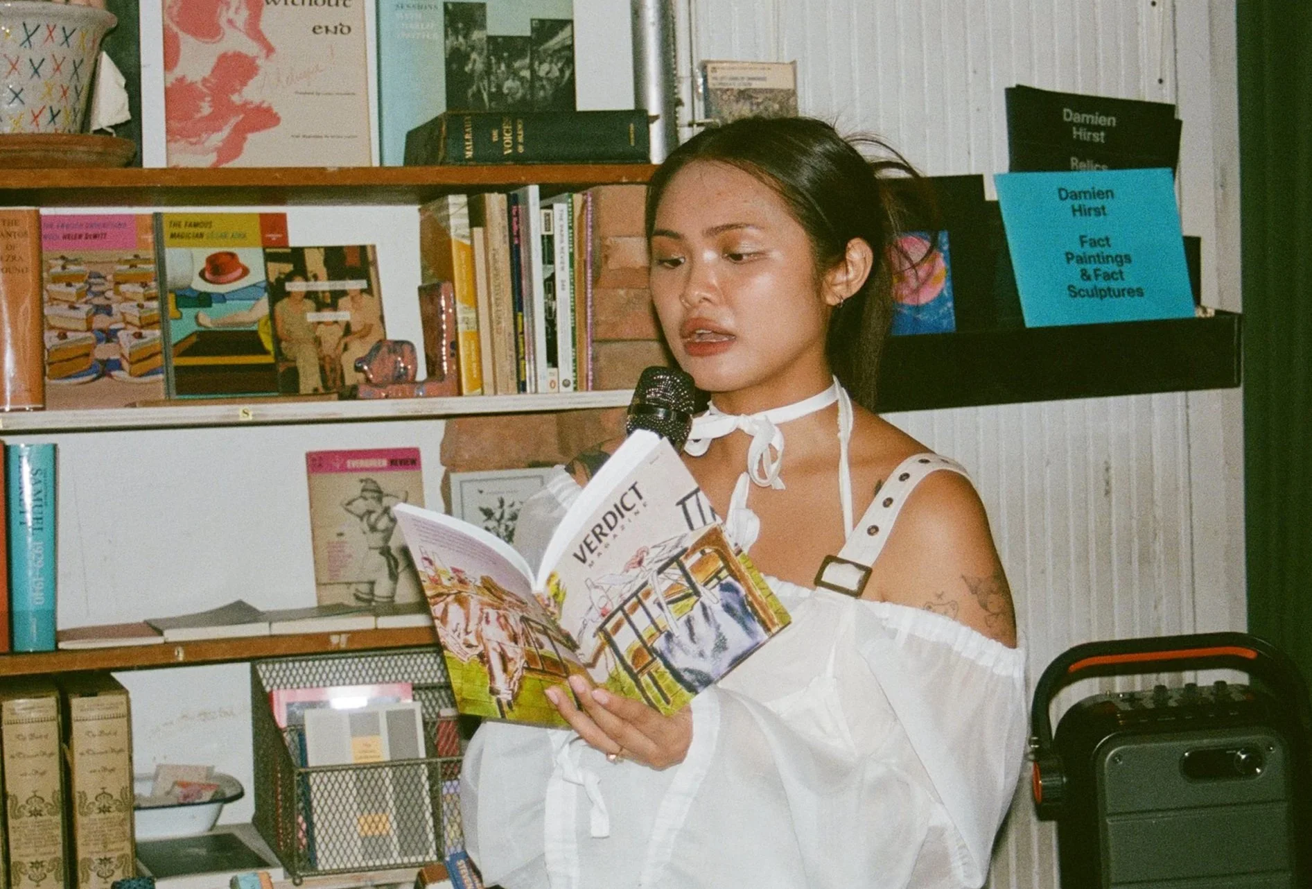 A woman reading a book aloud in a bookstore or library setting, holding a microphone.