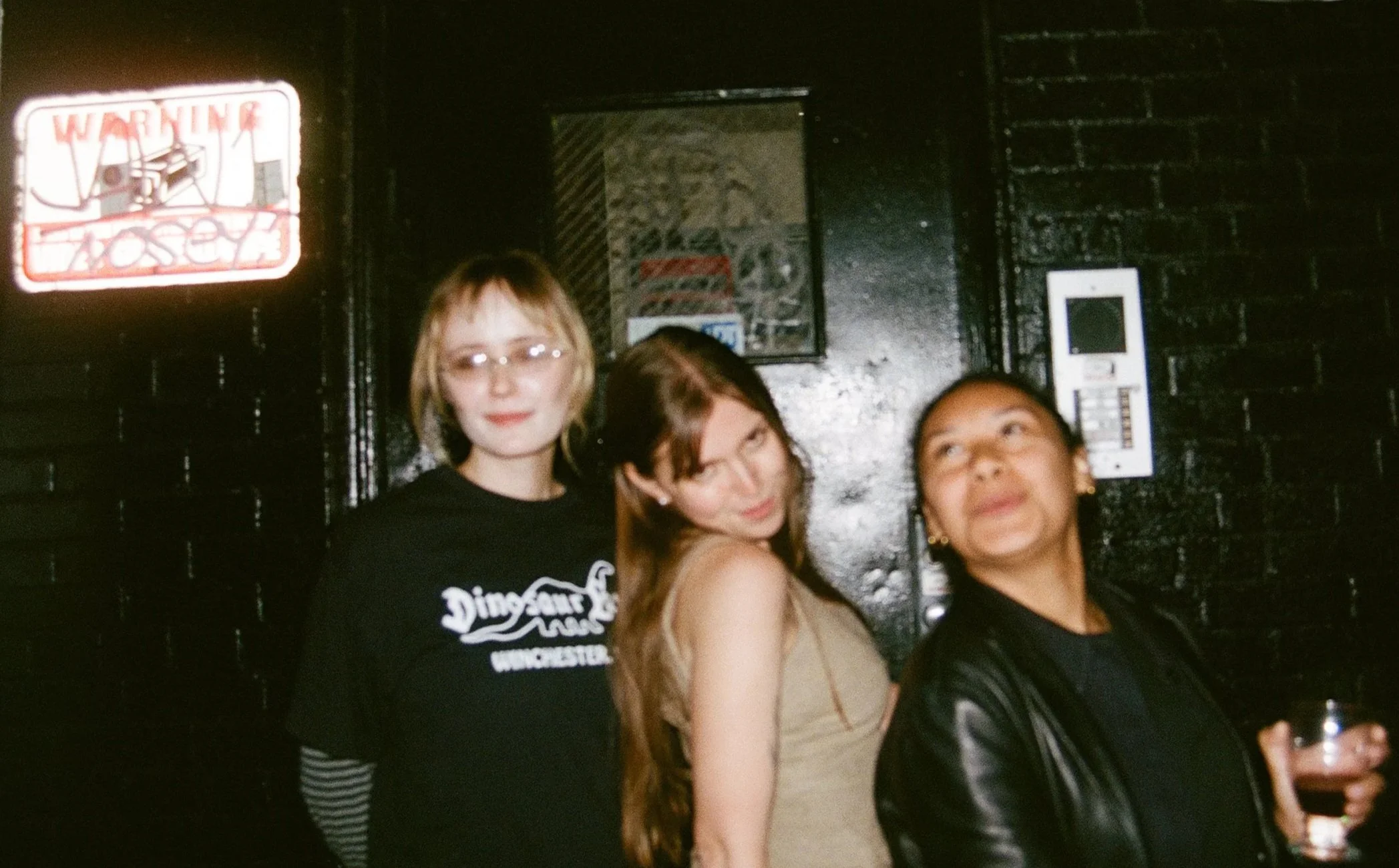 Three women standing in front of a black brick wall with neon signs, one holding a drink.
