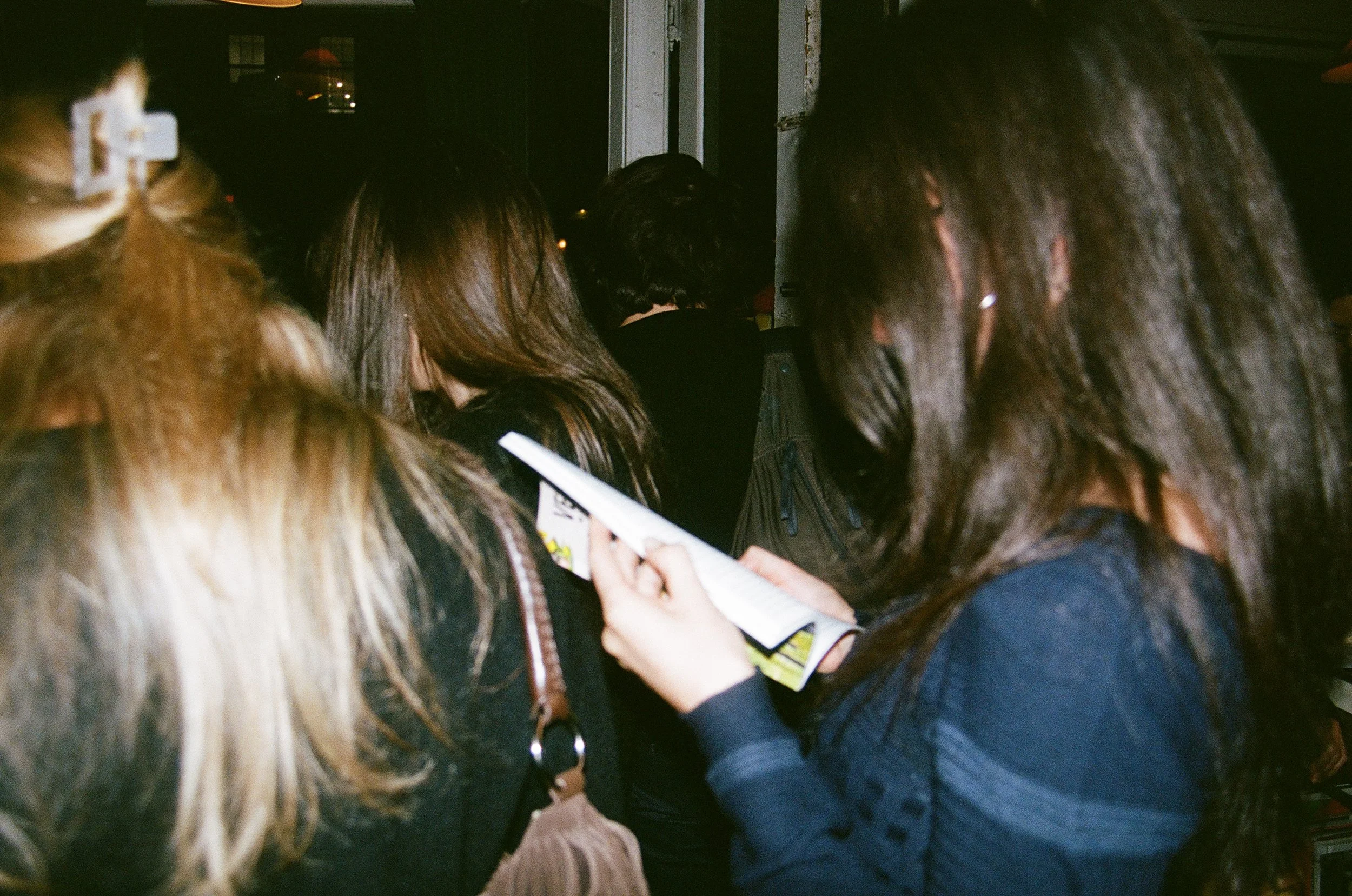 Group of women at a crowded indoor event, some looking at a paper or pamphlet.