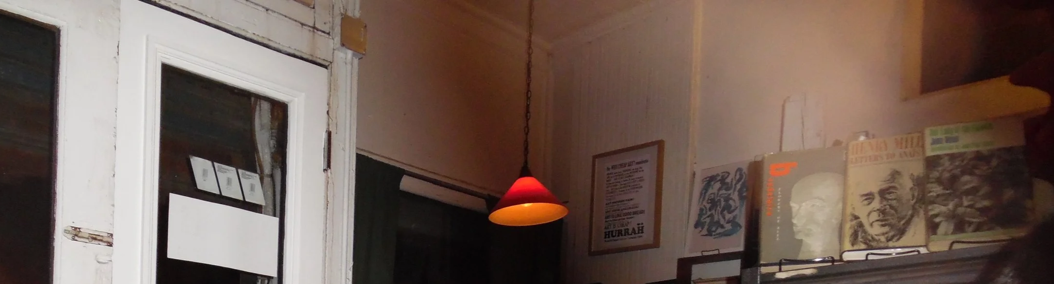 Interior of a room with a white door and window on the left, a hanging red lampshade in the center, and a shelf with books and framed pictures on the right wall.