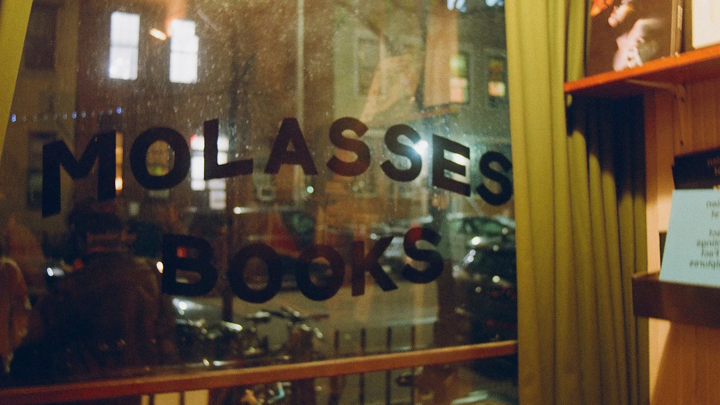 View through a window with the words 'MOLASSES BOOKS' written on it, showing parked cars and a building outside at night.