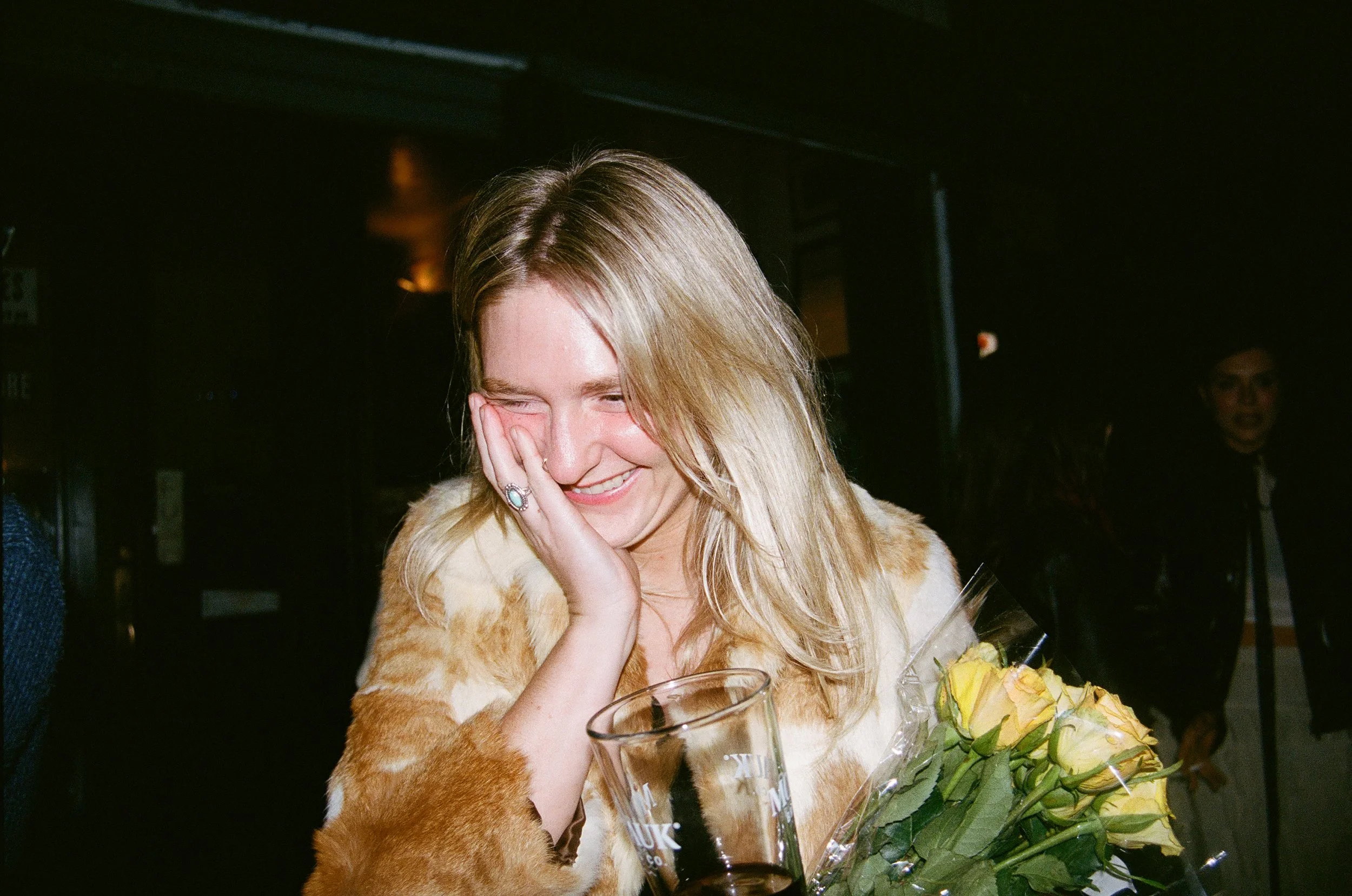 A young woman with blonde hair, smiling and covering her face with her hand, holding a bouquet of yellow roses, wearing a fur coat, in a dimly lit indoor setting.