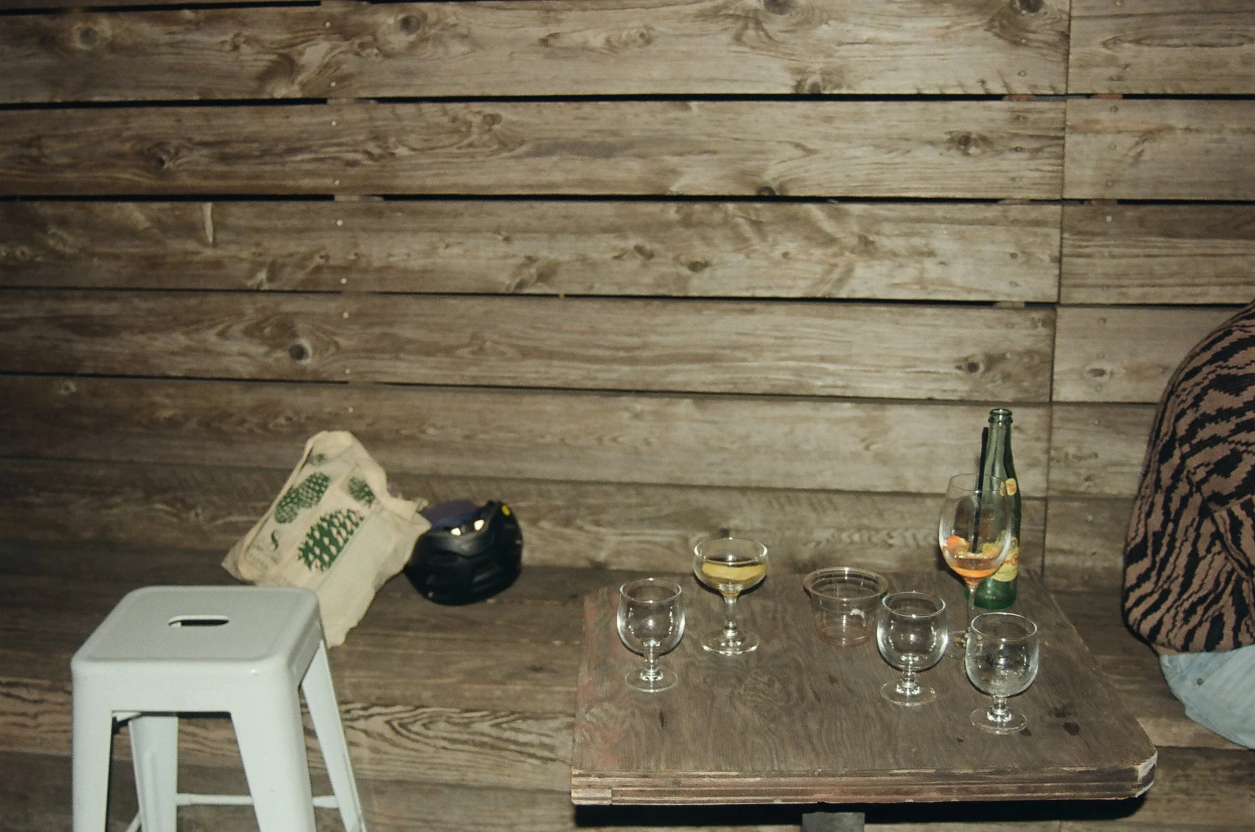 A wooden table with empty glasses and a couple of bottles, a stool, and a bag on a wooden bench against a wood-paneled wall.