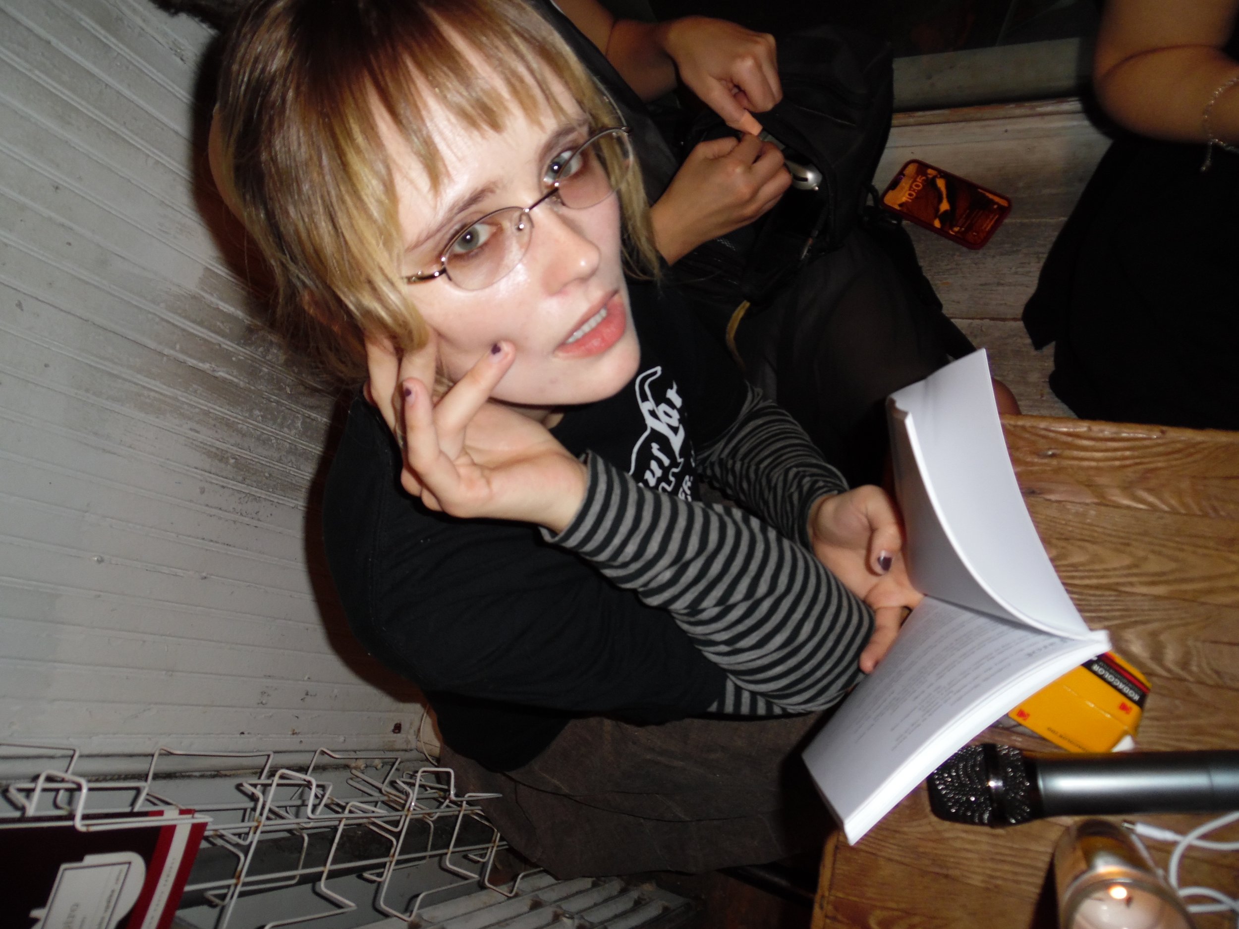 Young woman with glasses and striped sleeves resting her chin on her hand, looking up at the camera while sitting at a wooden table with an open book, with various items around her including a smartphone, backpack, and microphone.