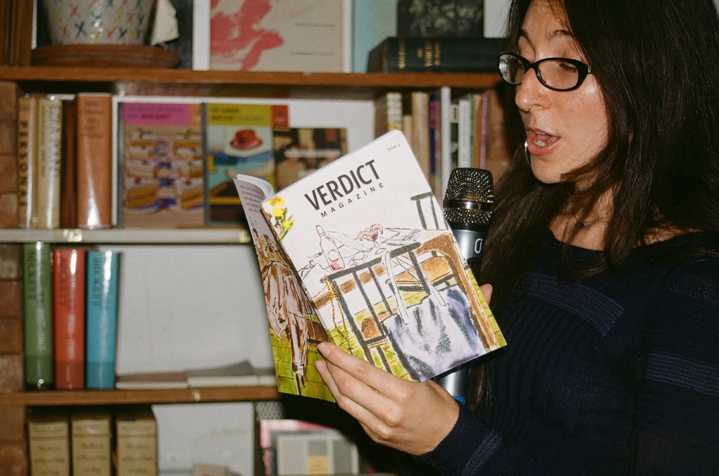 A woman with glasses reading a magazine titled "Verdict" while speaking into a microphone in a library.