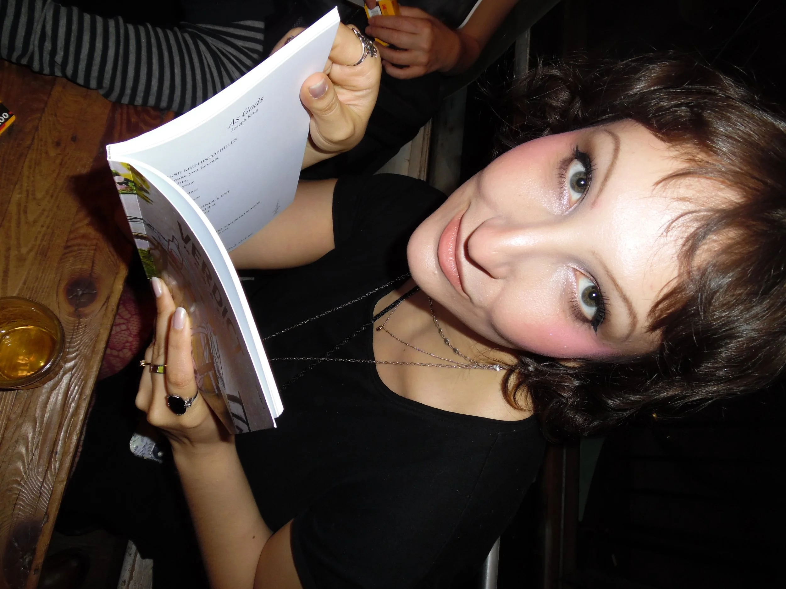 A woman with short brown hair and light eyes sitting at a wooden table, holding a book, with a glass of drink on the table.