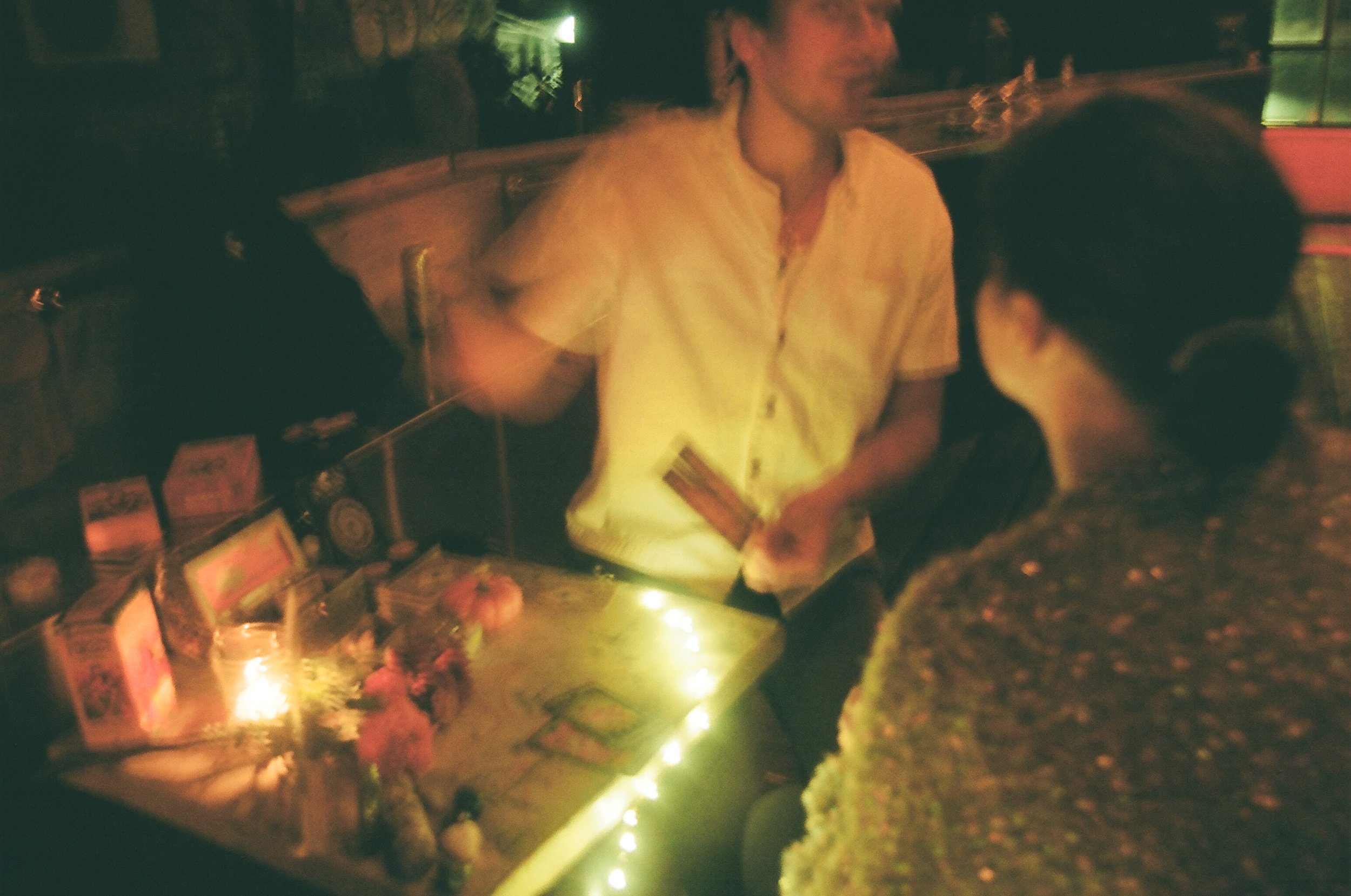 A person with long hair and a cream-colored shirt is standing at a table, talking to a woman with glasses and a dark patterned shirt inside a dimly lit restaurant or bar venue.