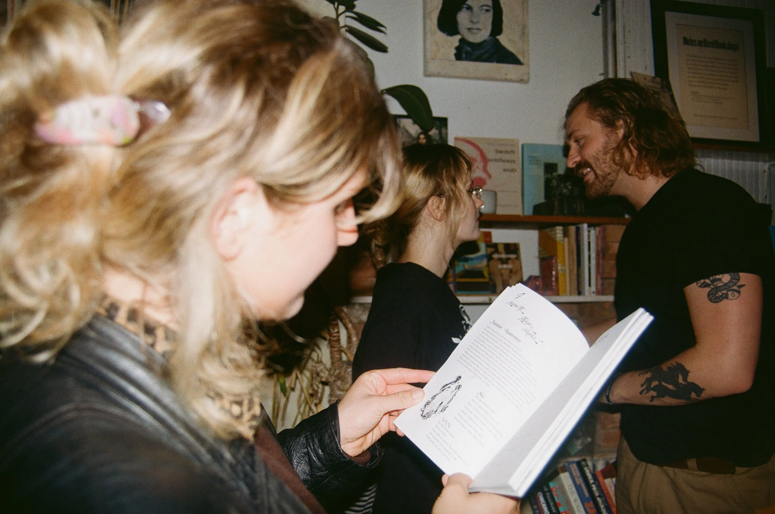 Three people in a bookstore or library, one woman with blond hair reading a book, woman with glasses in the background, man with curly hair and tattoos talking to her.