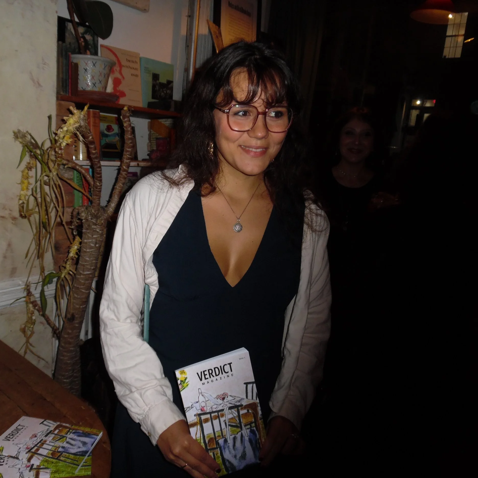 A woman with glasses, wavy hair, and a necklace holding a magazine labeled 'Verdict' at an indoor event, with books and a plant in the background.