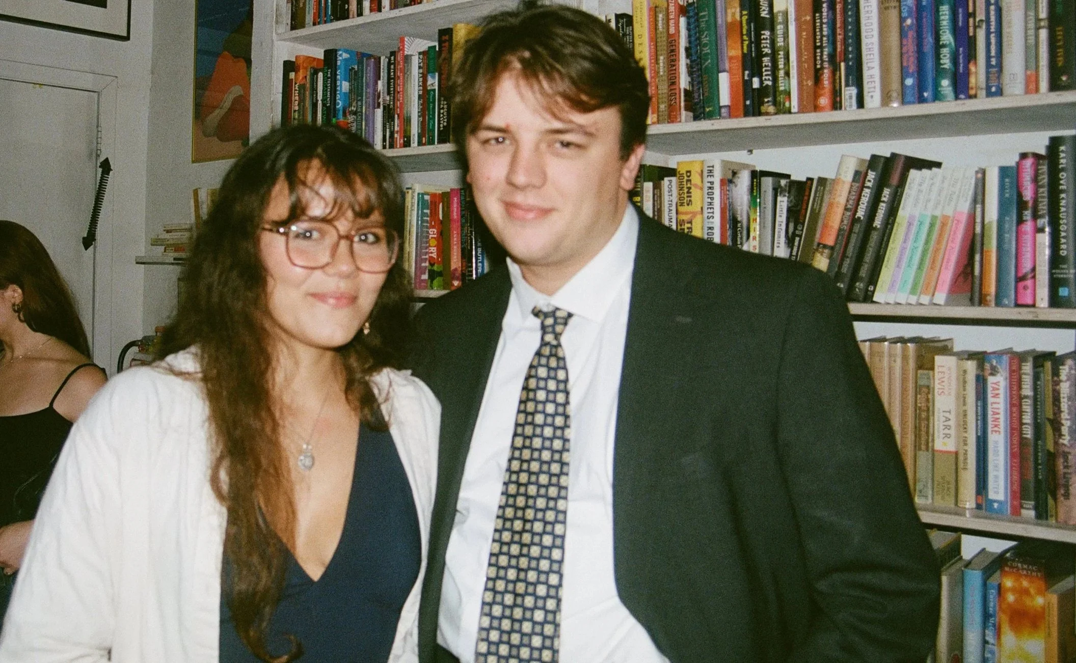 A woman with glasses and long brown hair, wearing a white blazer over a navy dress, standing next to a man in a black suit and patterned tie in front of a bookshelf filled with books at a social event.
