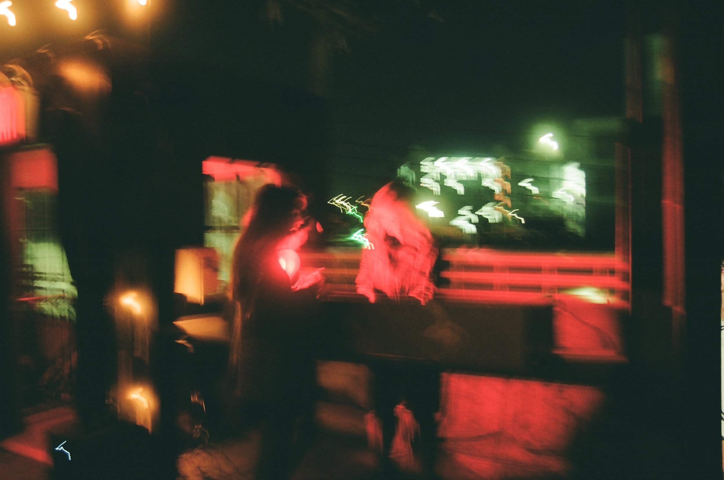 Blurry photo of two people standing indoors at night, illuminated by red and green neon lights.