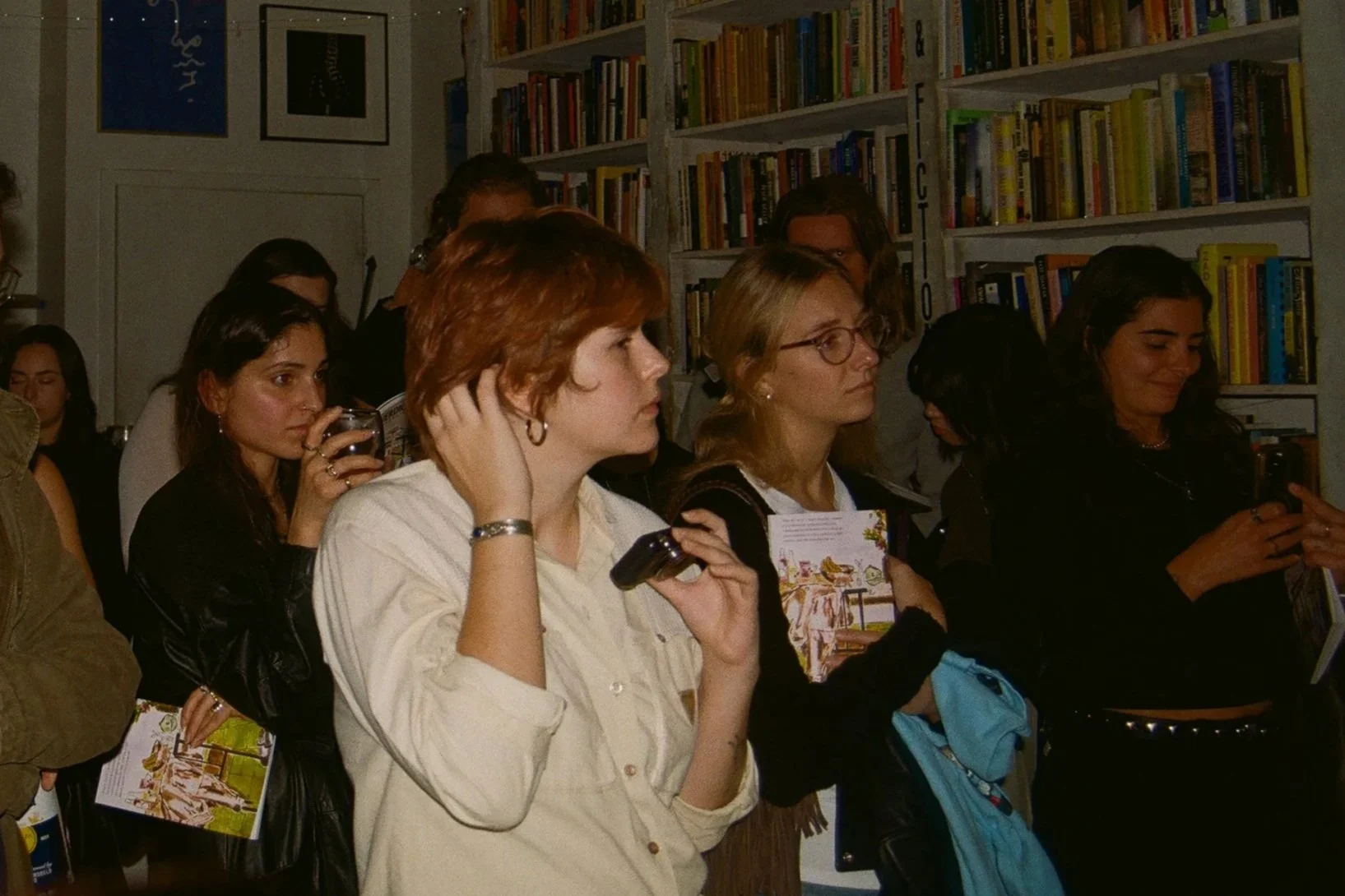 A group of women standing closely together, some holding brochures and one taking a photo with a smartphone, inside a bookstore with shelves filled with books in the background.