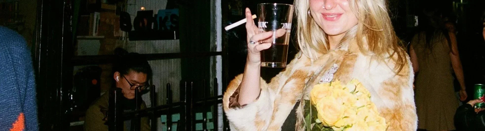 A woman with blonde hair wearing a furry coat holding a glass and a bouquet of yellow roses, smiling at a party or social gathering.