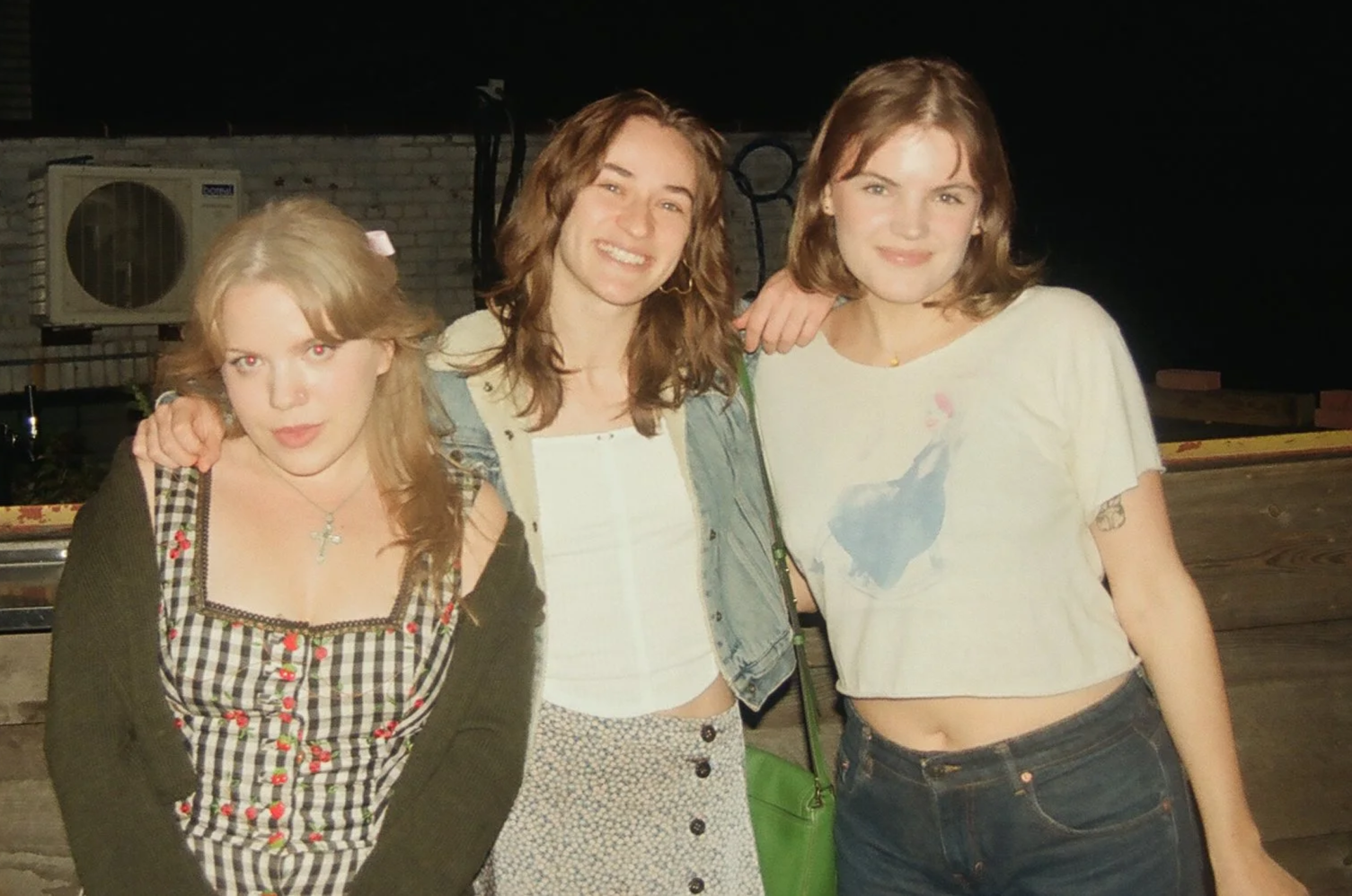 Three young women posing together outside at night, smiling, with a brick wall, air conditioning unit, and parking lot in the background.