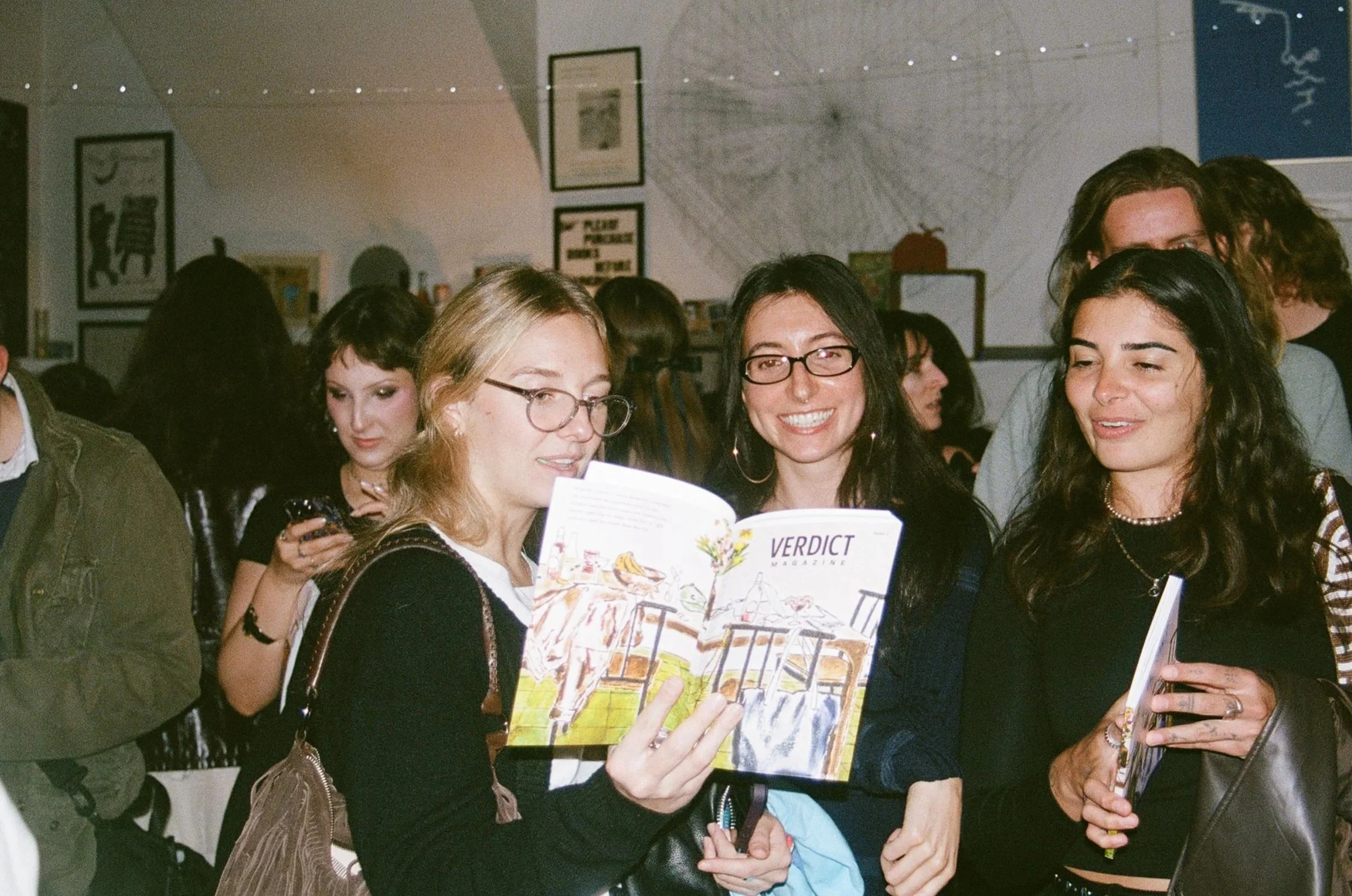 Three women are smiling and looking at a magazine titled 'Verdict' in a crowded indoor setting, possibly a bookstore or gallery.
