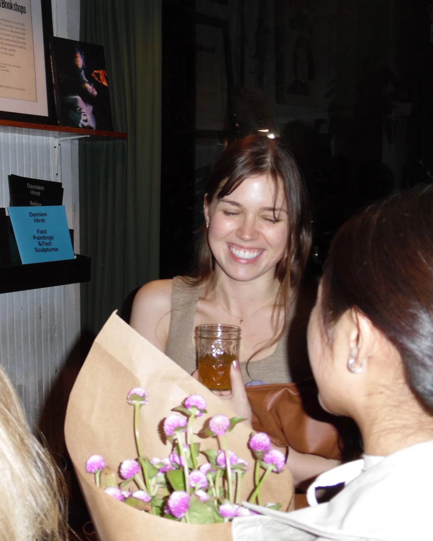 A woman is smiling with eyes closed, holding a glass of amber-colored beverage, and receiving a bouquet of pink flowers wrapped in brown paper from a person with dark hair and earrings.