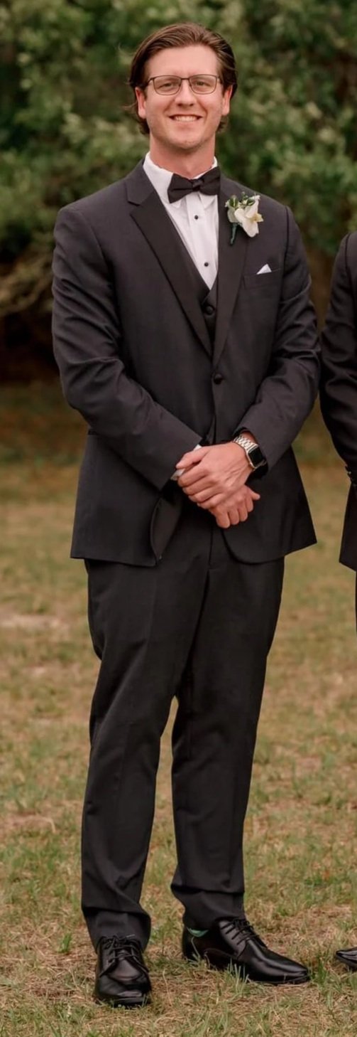 Man in black tuxedo with bow tie and boutonniere, standing outdoors on grass.