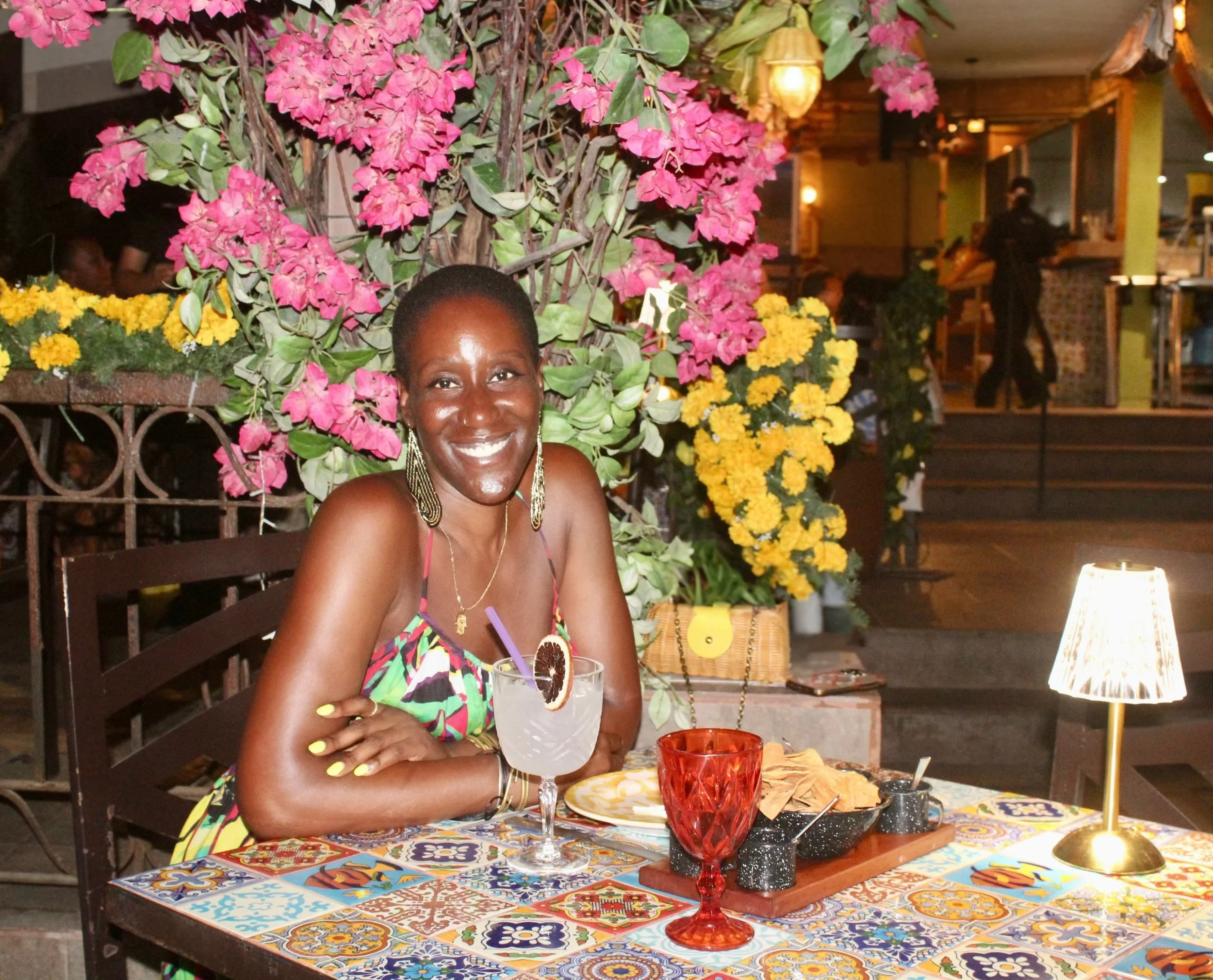 Smiling woman sitting at a table in a restaurant with pink and yellow flowers behind her, holding a cocktail with a slice of blood orange, at night.