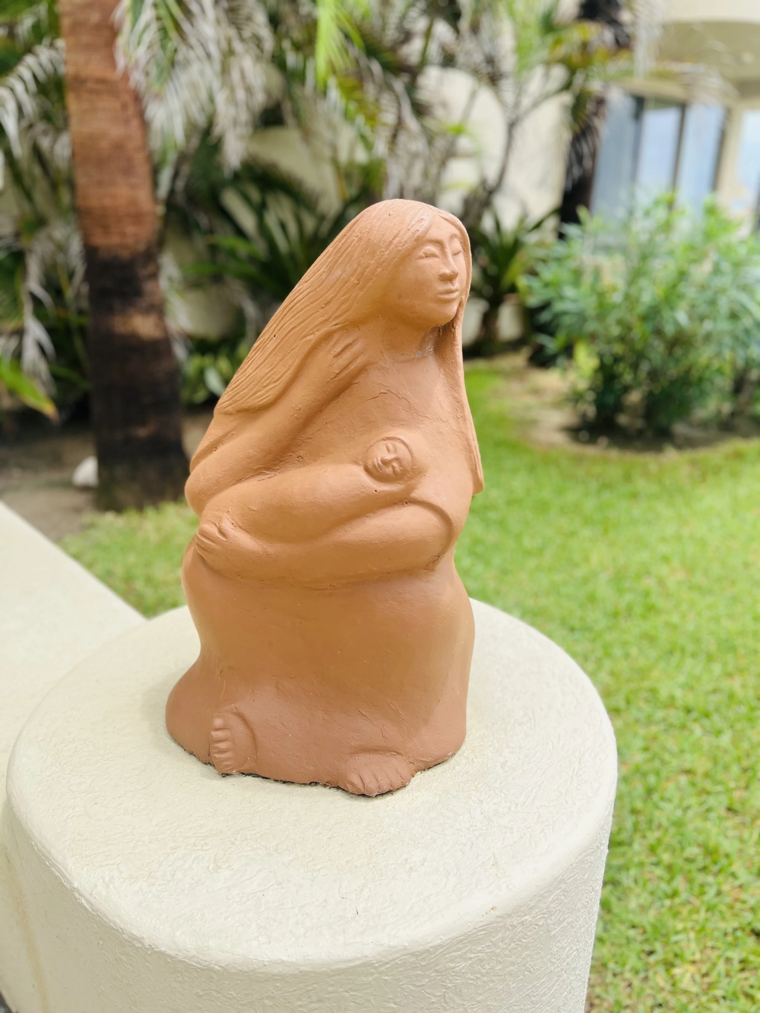 Clay sculpture of a pregnant woman with long hair, standing outdoors in a garden setting.