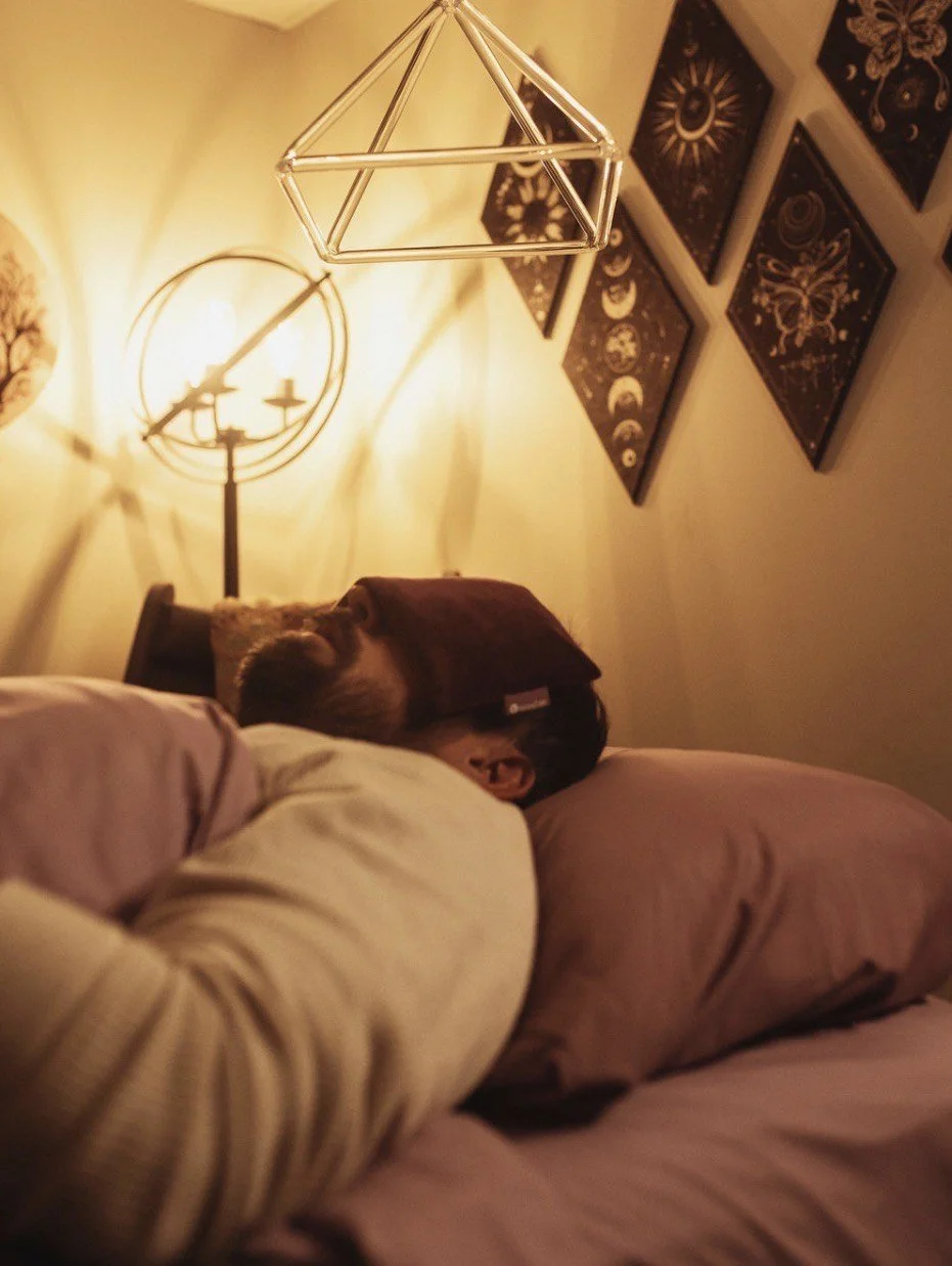 Person sleeping in bed with a blanket covering their face, a pillow, and warm lighting from a lamp and a geometric light fixture on the ceiling.