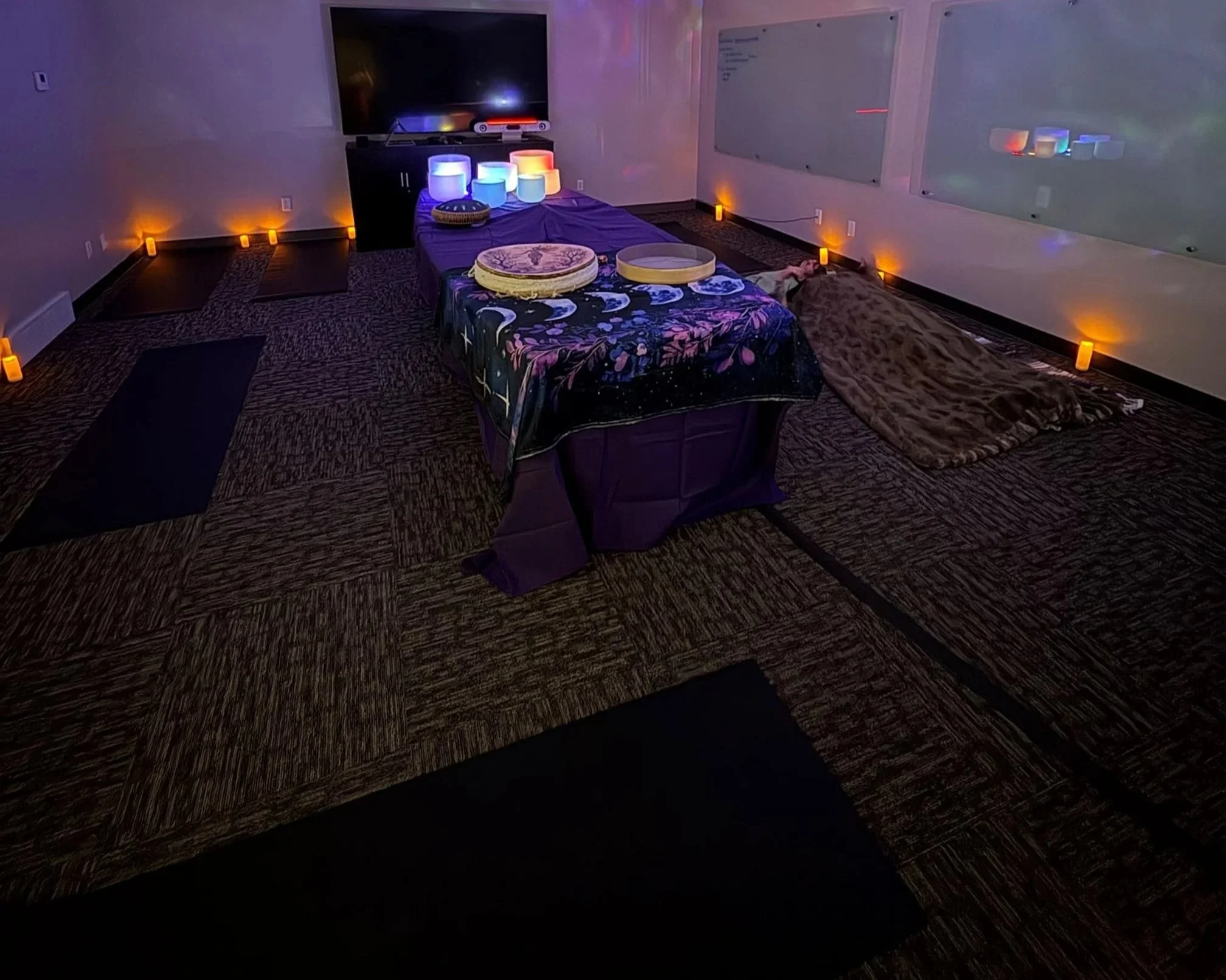 A dimly lit room with candles around the perimeter, a large flat-screen TV, and yoga mats on the patterned carpet. In the center, there are tables with massage or therapy tools, and a person lying on a furry blanket on the floor.