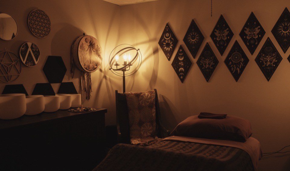 A cozy room with warm lighting, decorated with black hexagonal and diamond-shaped wall art. A bed with pillows and folded blankets, a tall chair, and a standing lamp with three candles inside create a relaxing ambiance.