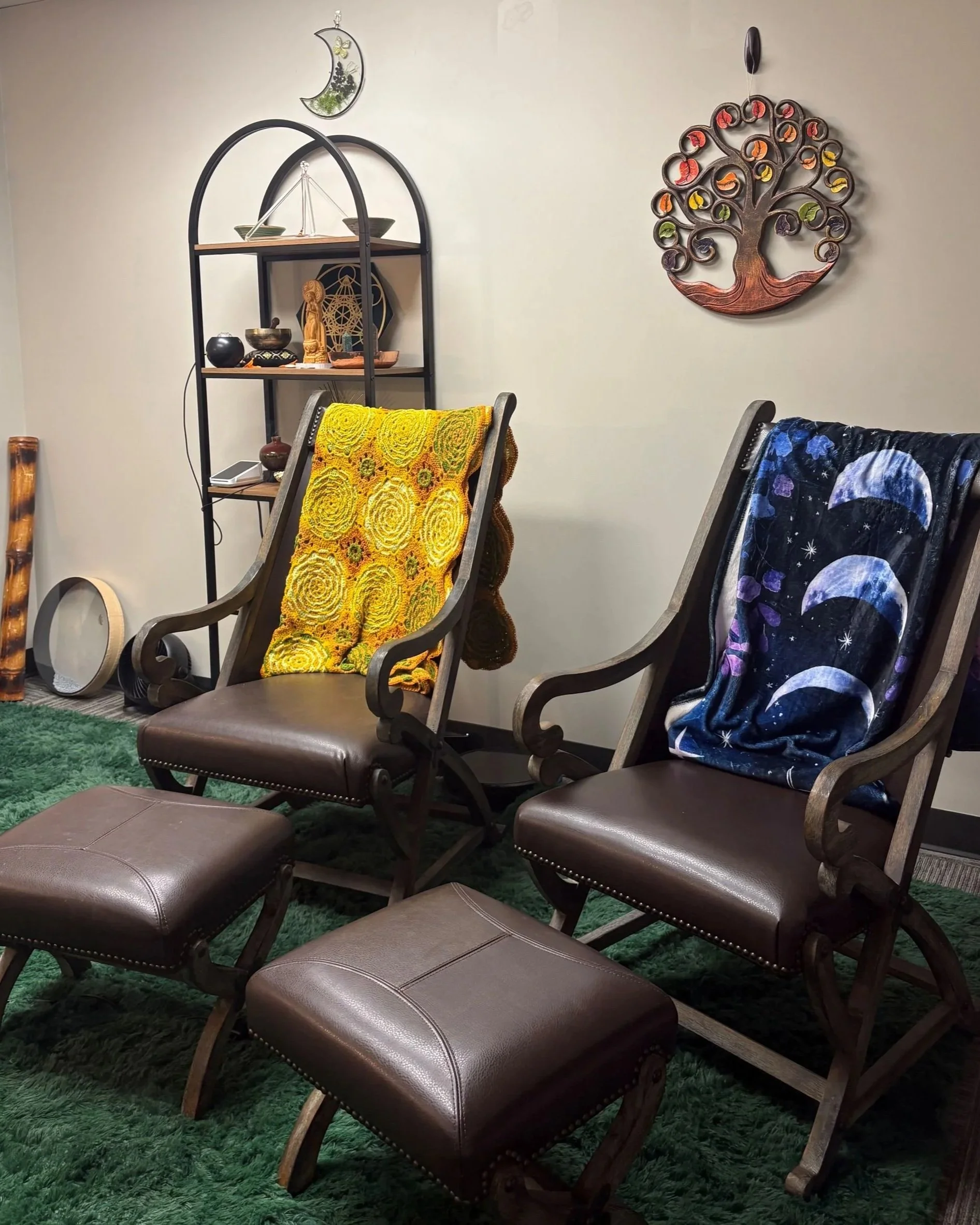 Living room with two wooden chairs with tufted leather cushions, each draped with a colorful blanket, and two matching ottomans. A green shag rug covers the floor, and a white wall with decorative wall art, including a tree sculpture and a moon-shaped ornament, is visible. There is a black metal shelf holding various decorative items.