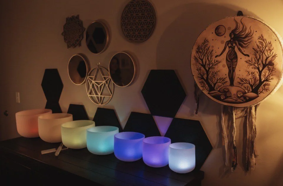 A collection of six glowing singing bowls in pastel colors, arranged on a surface against a wall decorated with various geometric and mirror wall art, with a large round artwork featuring a woman standing between trees illuminated by soft yellow ligh