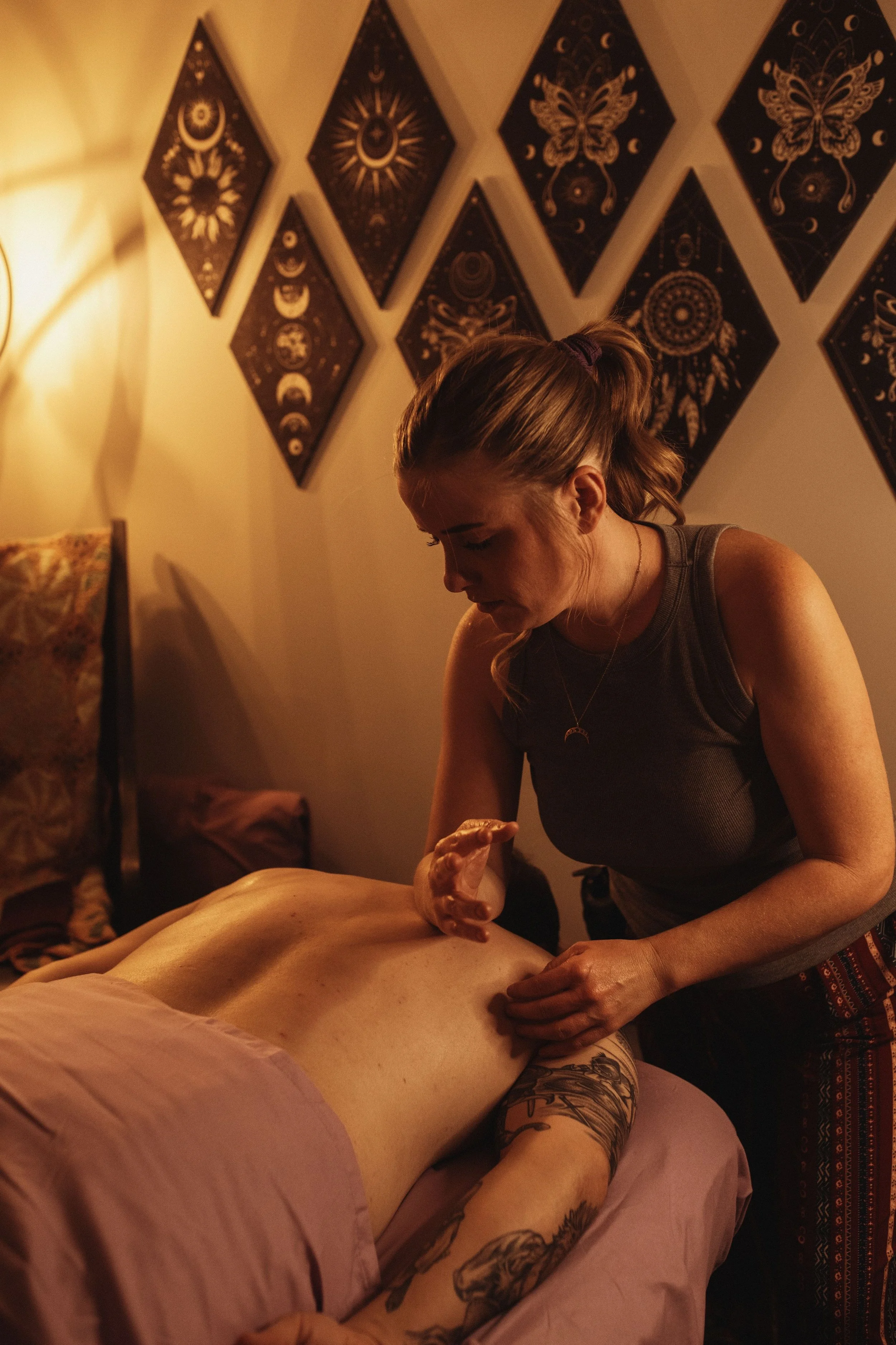 A woman giving a back massage to a man lying face down on a massage table in a dimly lit room with dark wall art.