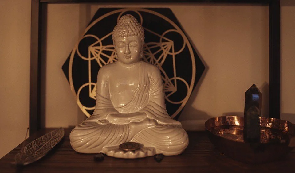 A white Buddha statue seated in a meditative pose with a decorative geometric artwork behind it, surrounded by an oval tray and a black gemstone on a wooden surface.