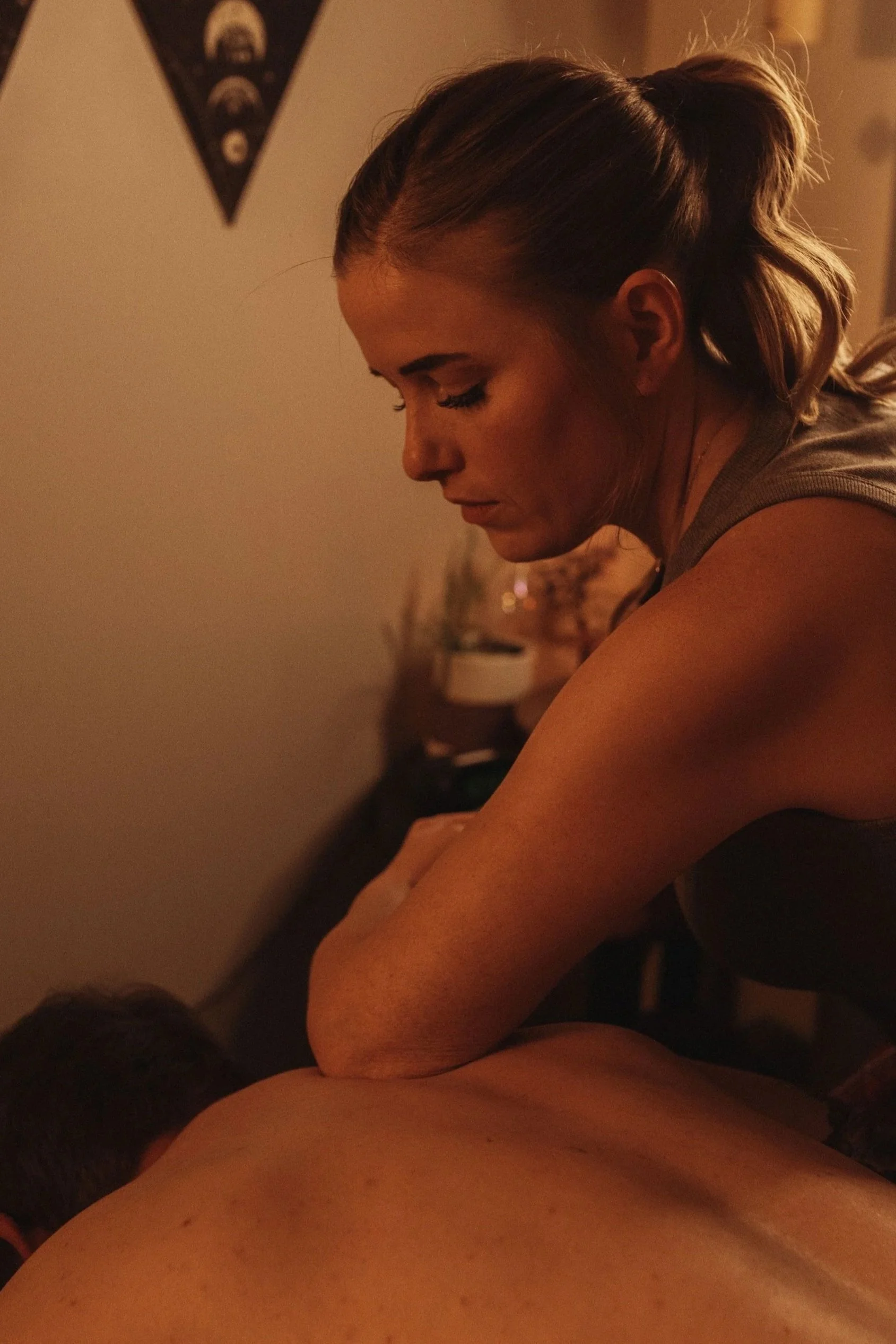 A woman with blonde hair in a ponytail giving a massage to a man lying face down, in a dimly lit room.