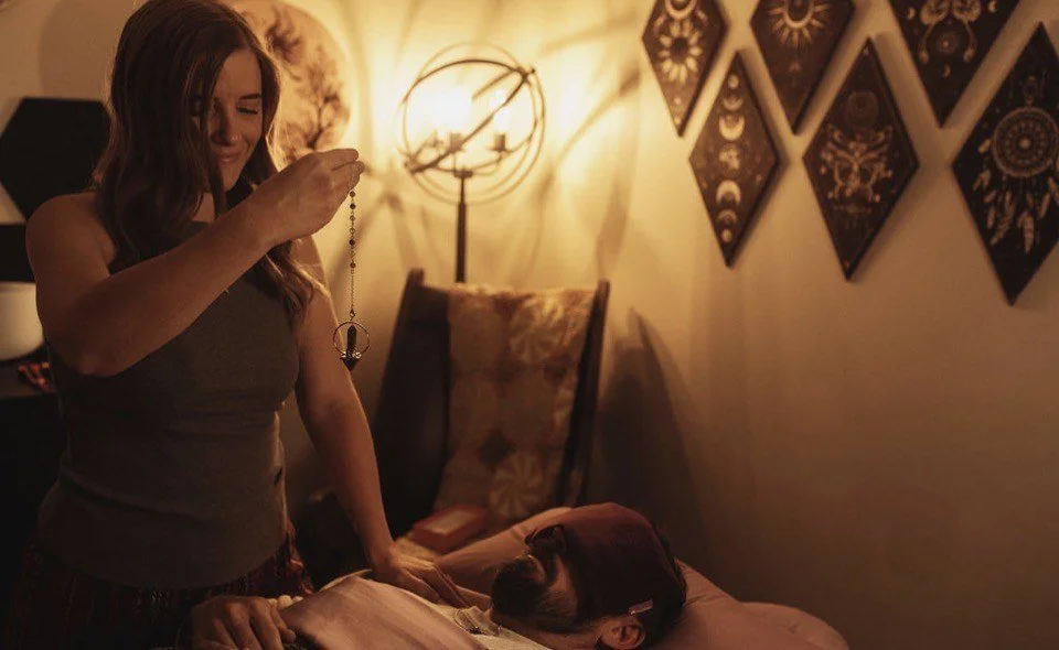 A woman standing in a dimly lit room holding a dreamcatcher, with a man laying in bed beside her, under warm lighting and decorated with wall art.
