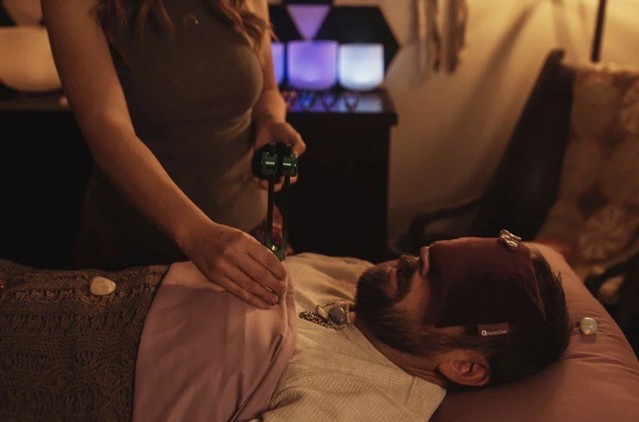 A woman giving a crystal healing or energy cleansing session to a man lying on a bed in a dimly lit room with colorful lights in the background.