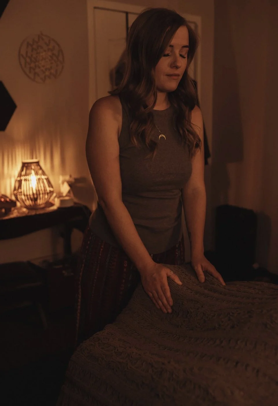 A woman standing at a table in a dimly lit room with warm lighting, eyes closed, wearing a sleeveless top and patterned pants.