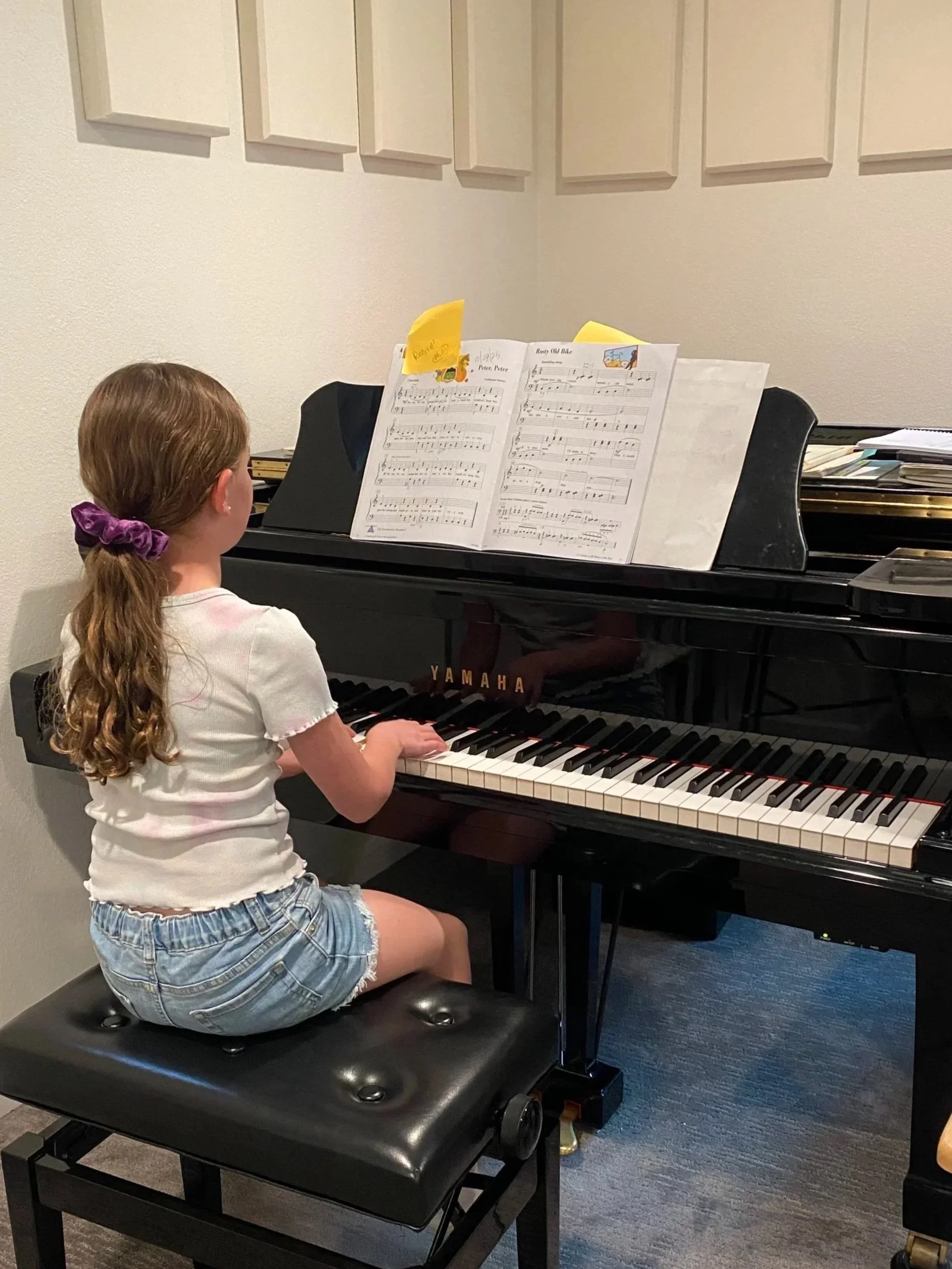A young girl with long curly hair, wearing a purple scrunchie, a white t-shirt, and denim shorts, sitting at a black Yamaha piano playing with sheet music in front of her.