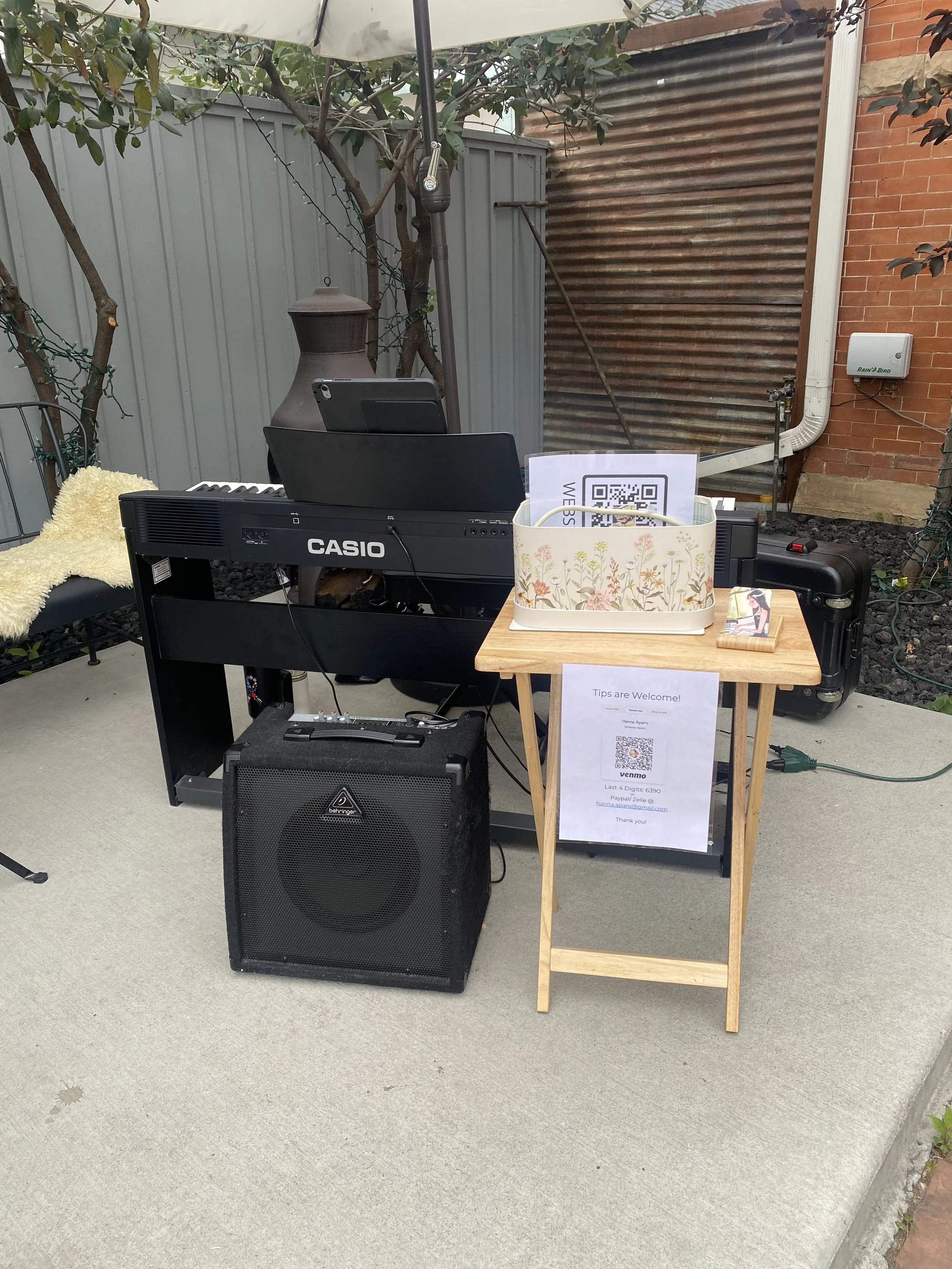 Outdoor setup with a keyboard, tablet, and speaker on a patio, with a wooden table holding signs with QR codes and a small framed photo, surrounded by trees, a fence, and a brick wall.