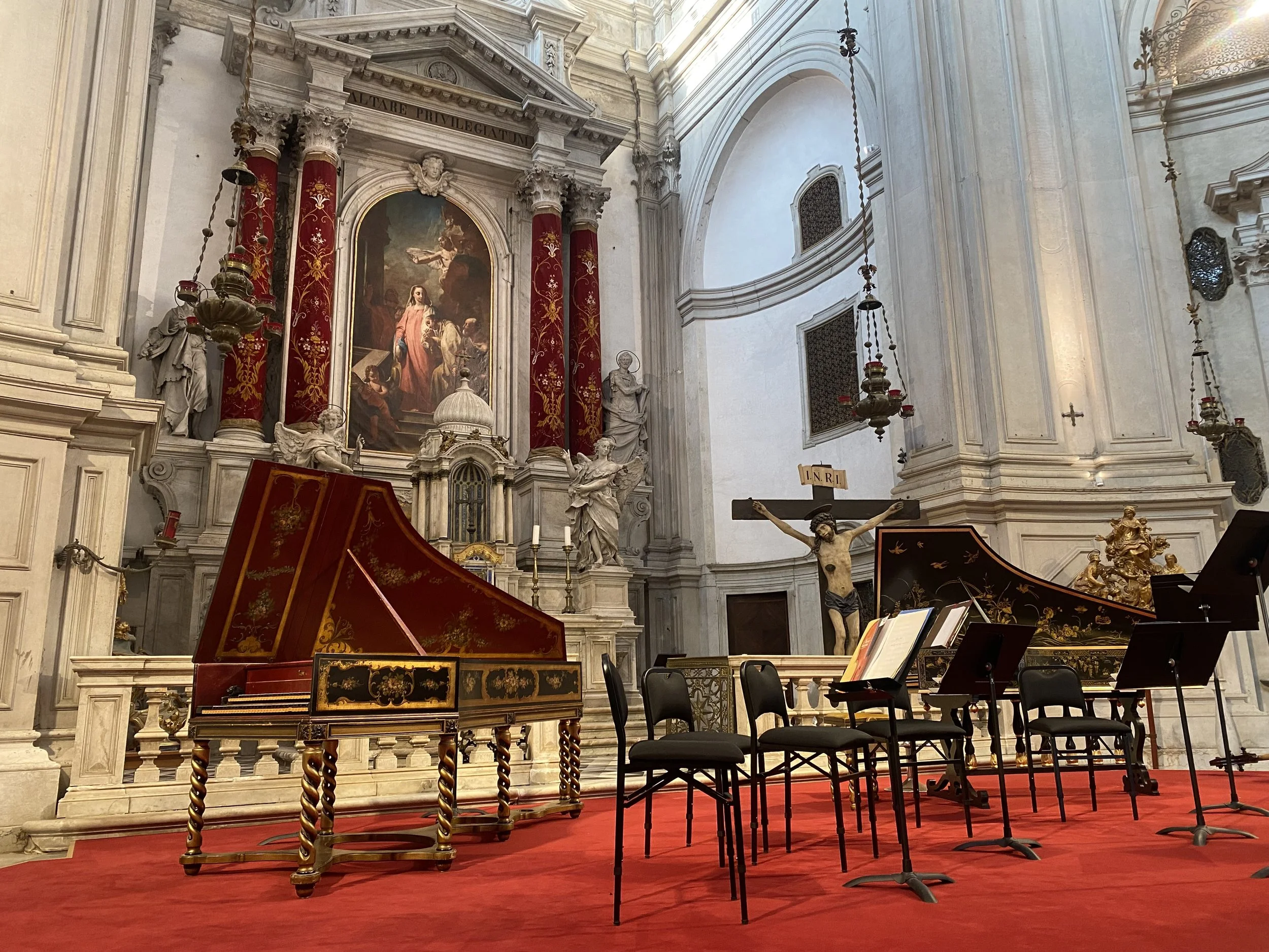 Inside a church with ornate walls, art, and sculptures, featuring a painted altar, a crucifix with Jesus, a harpsichord, music stands with sheet music, and chairs on a red carpet.