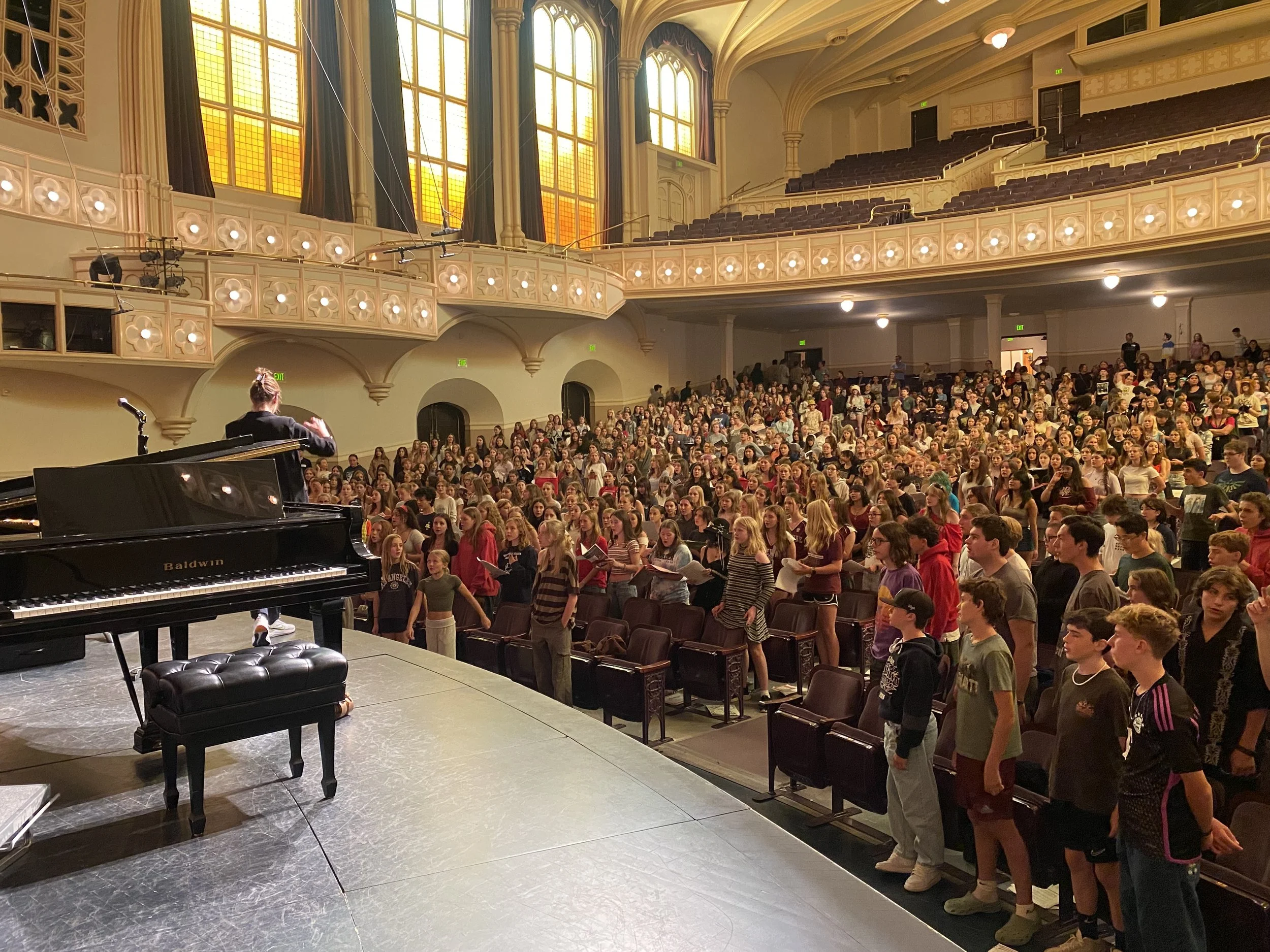 Large auditorium filled with an audience, with a person conducting in front of a grand piano on stage.