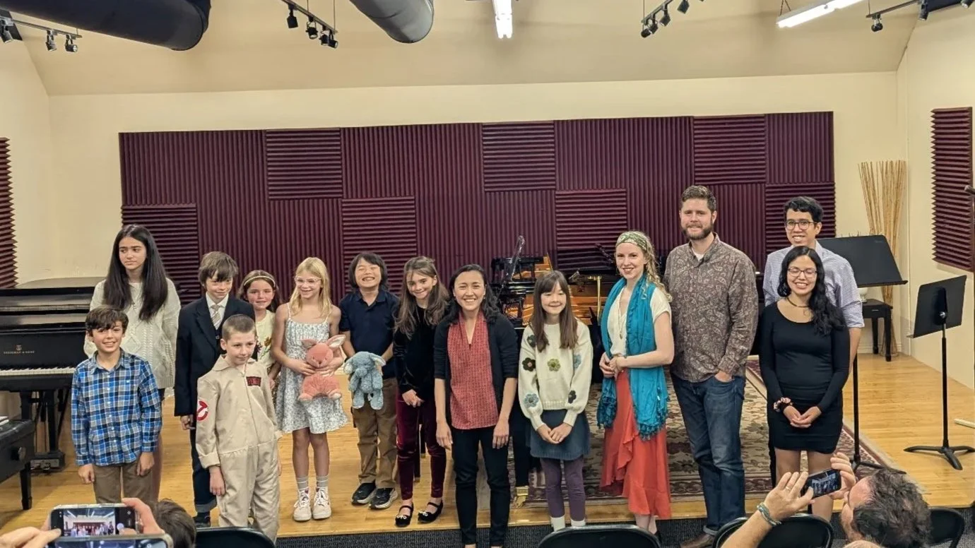 Group of children and adults standing on stage in a music classroom, with pianos and soundproofing panels in the background.