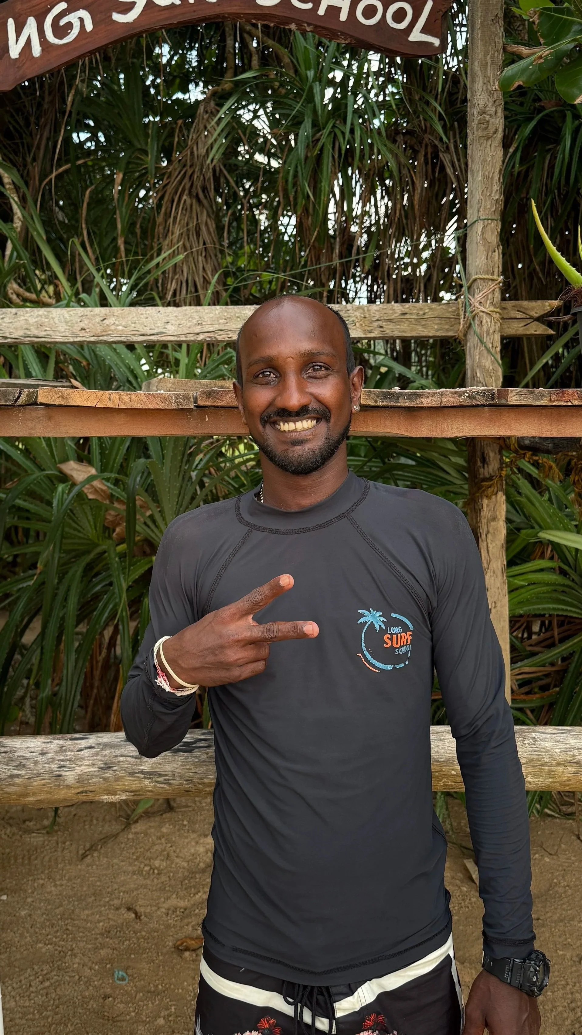 A smiling man with a dark shirt that reads 'Long Surf School' standing outdoors in front of lush green plants. He is making a peace sign with his right hand, wearing bracelets and a wristwatch, with a wooden sign in the background partially visible.