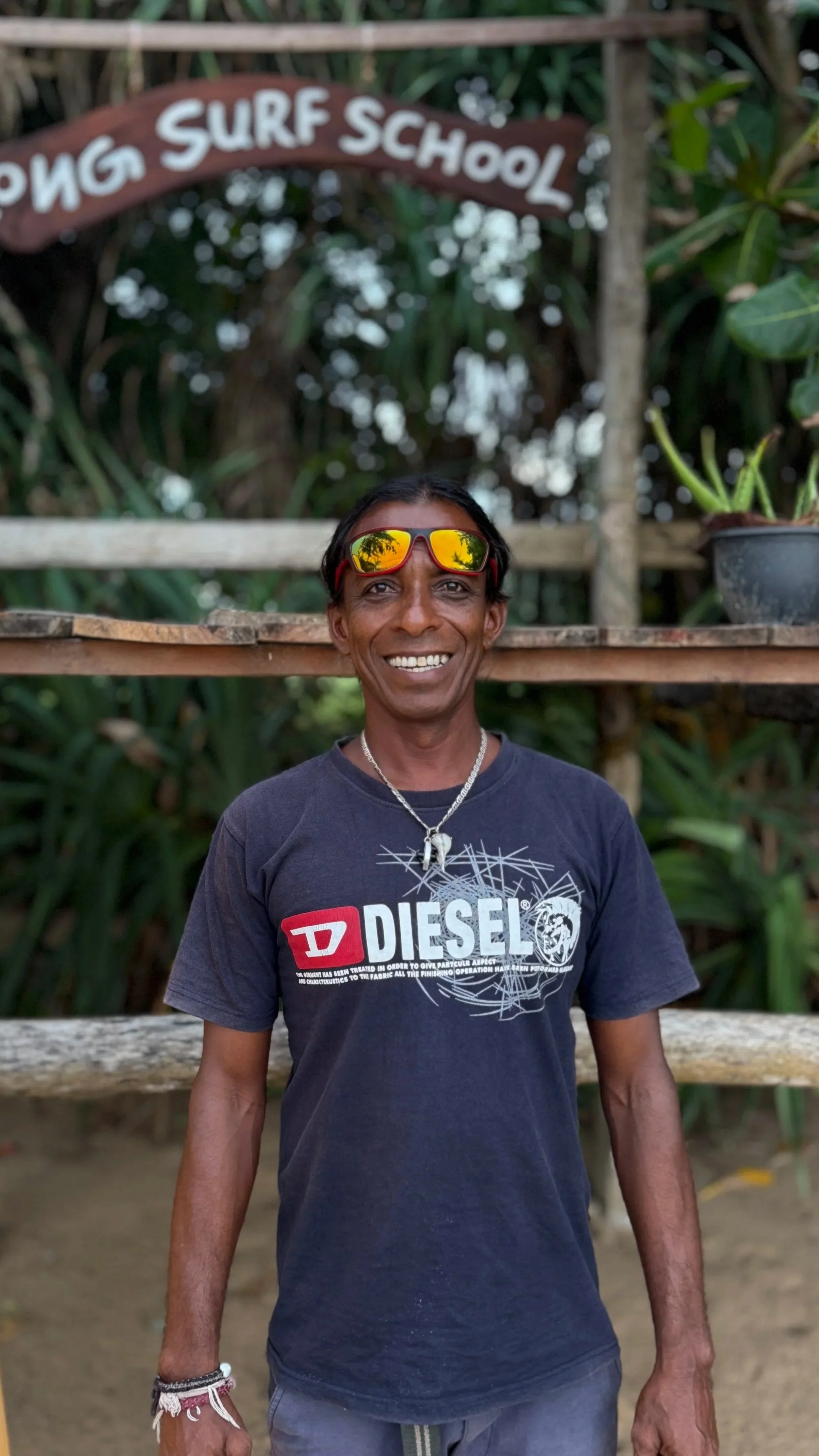 A smiling man wearing sunglasses, a Diesel t-shirt, and a necklace standing outdoors with a wooden surf school sign and green foliage in the background.